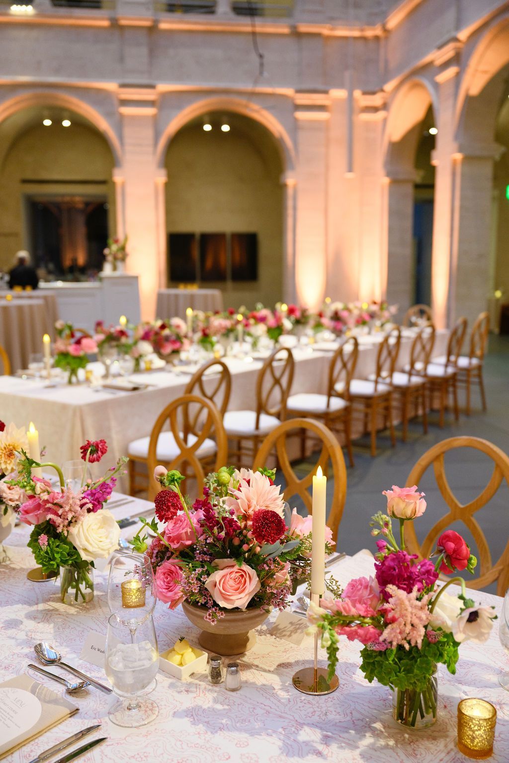 Long table set for an event with floral centerpieces, candles, and chairs in a venue with arches and warm lighting.