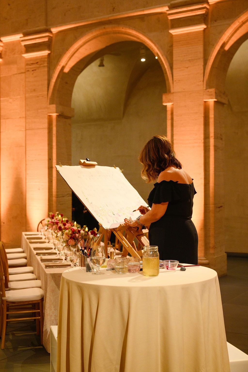 Woman painting at an easel in an arched hall, with a long table set with flowers and supplies.