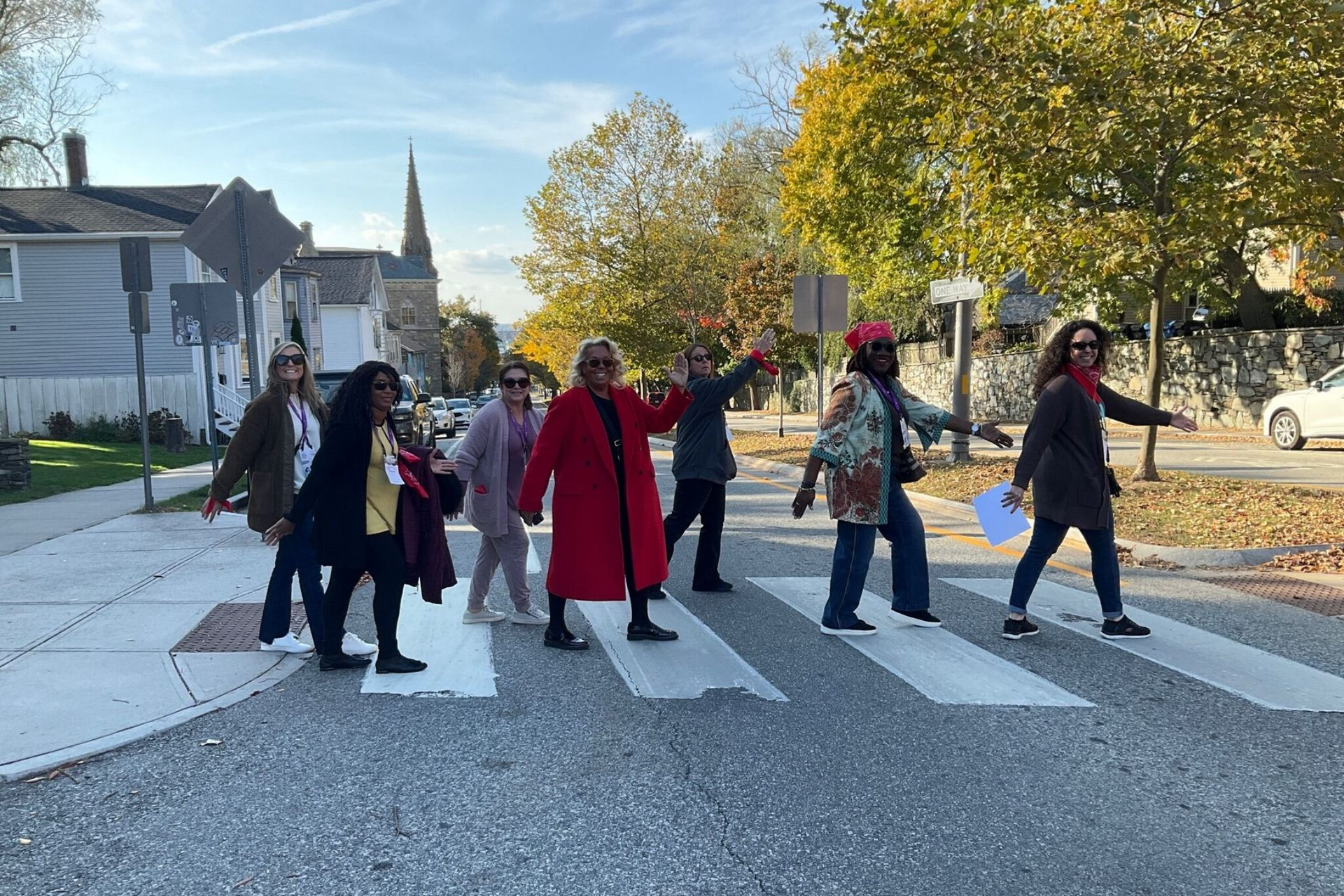 Group of people crossing a crosswalk on a city street; trees with fall foliage in the background.