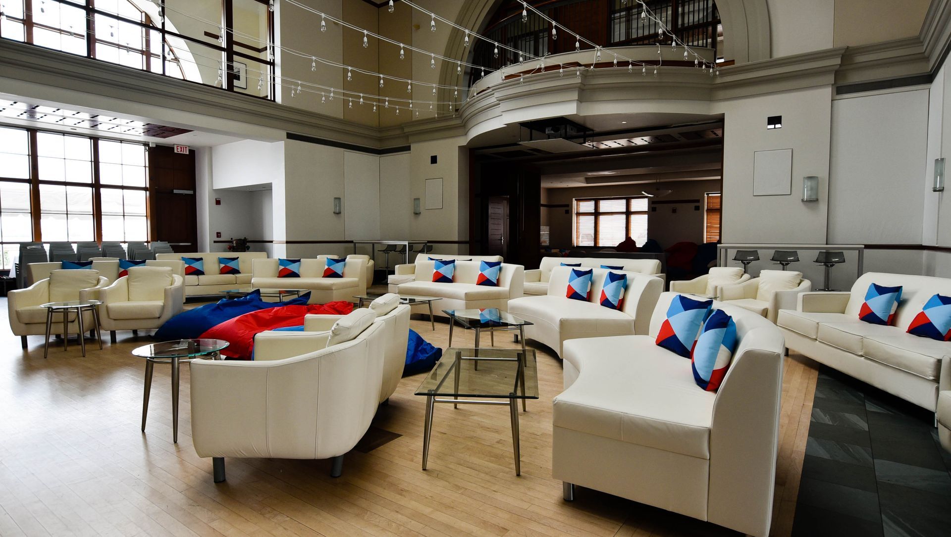 Lounge area with white couches, glass tables, and blue/red pillows. Large windows and balcony overhead.
