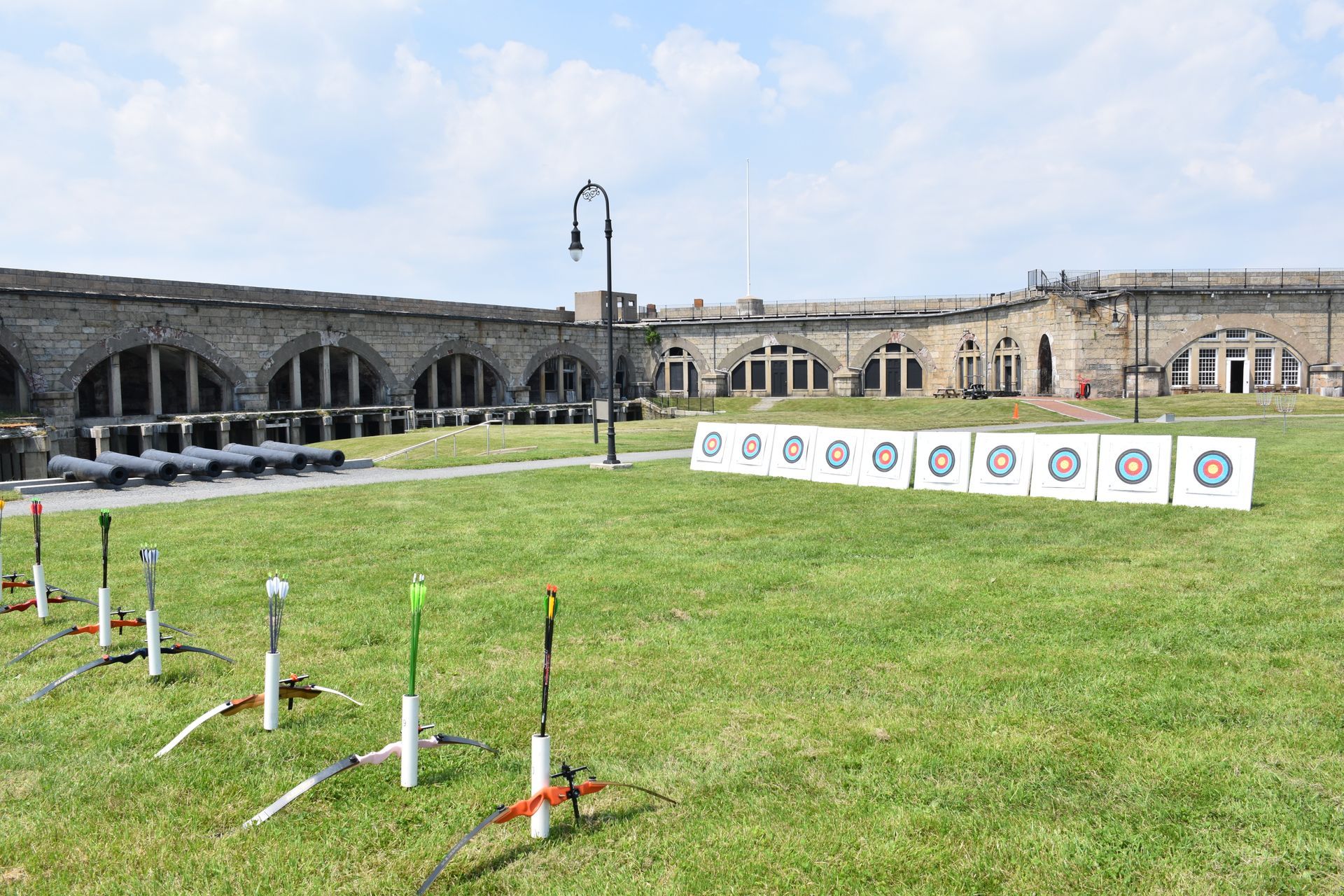 Archery targets on a grassy field, bows set out. A stone building with arched windows is in the background.