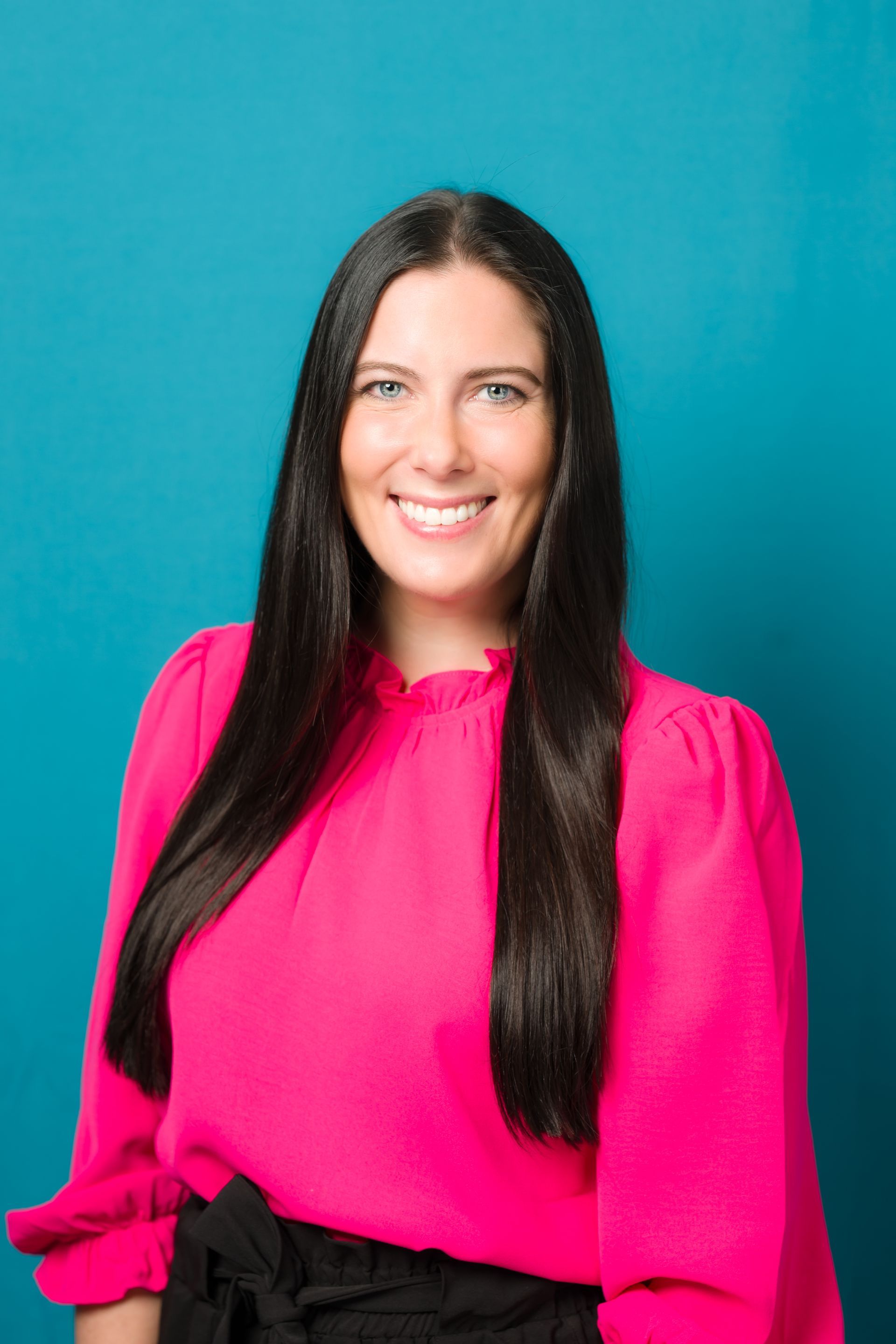 Woman with long, dark hair, wearing a pink blouse and smiling in front of a blue background.
