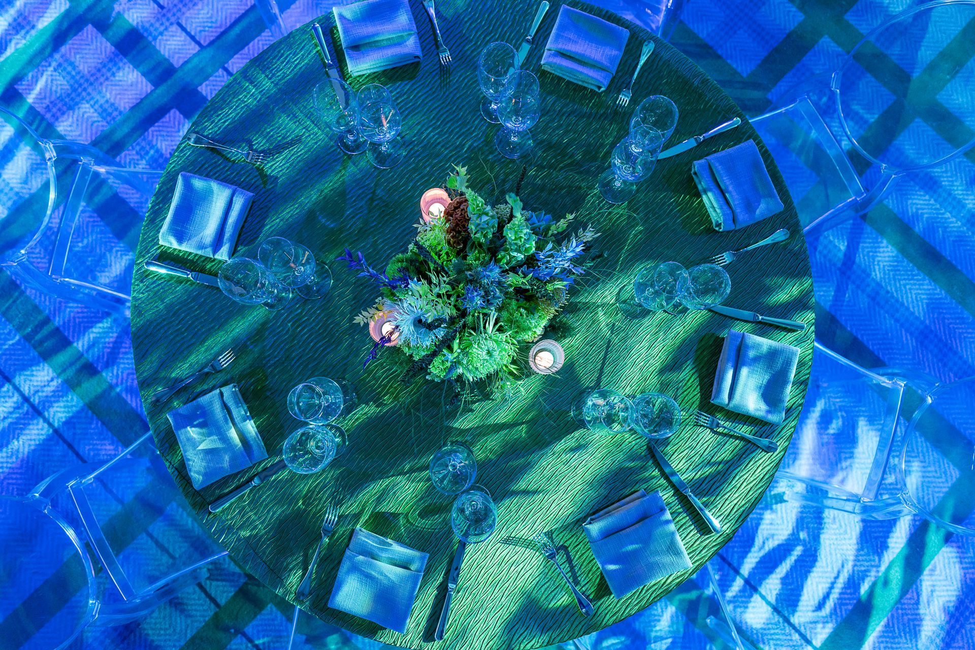A round table set for a gathering with blue and green accents, floral centerpiece, and clear settings.