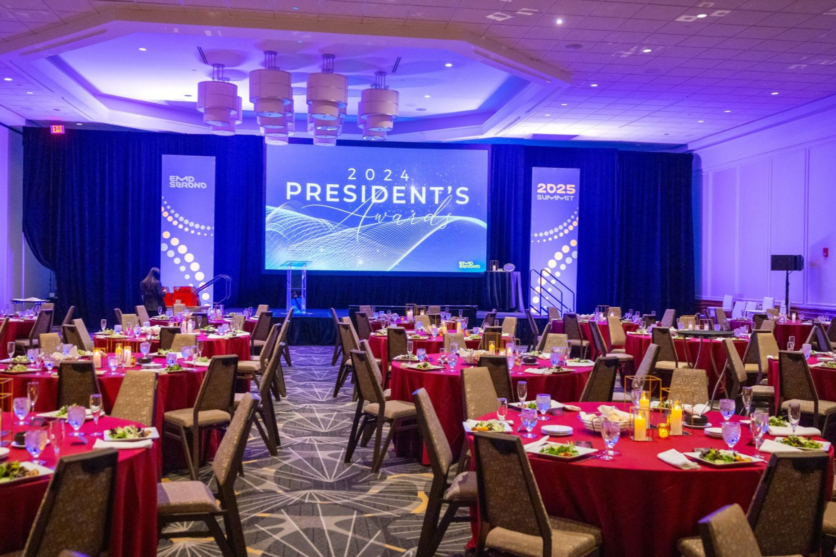 Large banquet hall set up for an event, red tablecloths, stage with screen reading