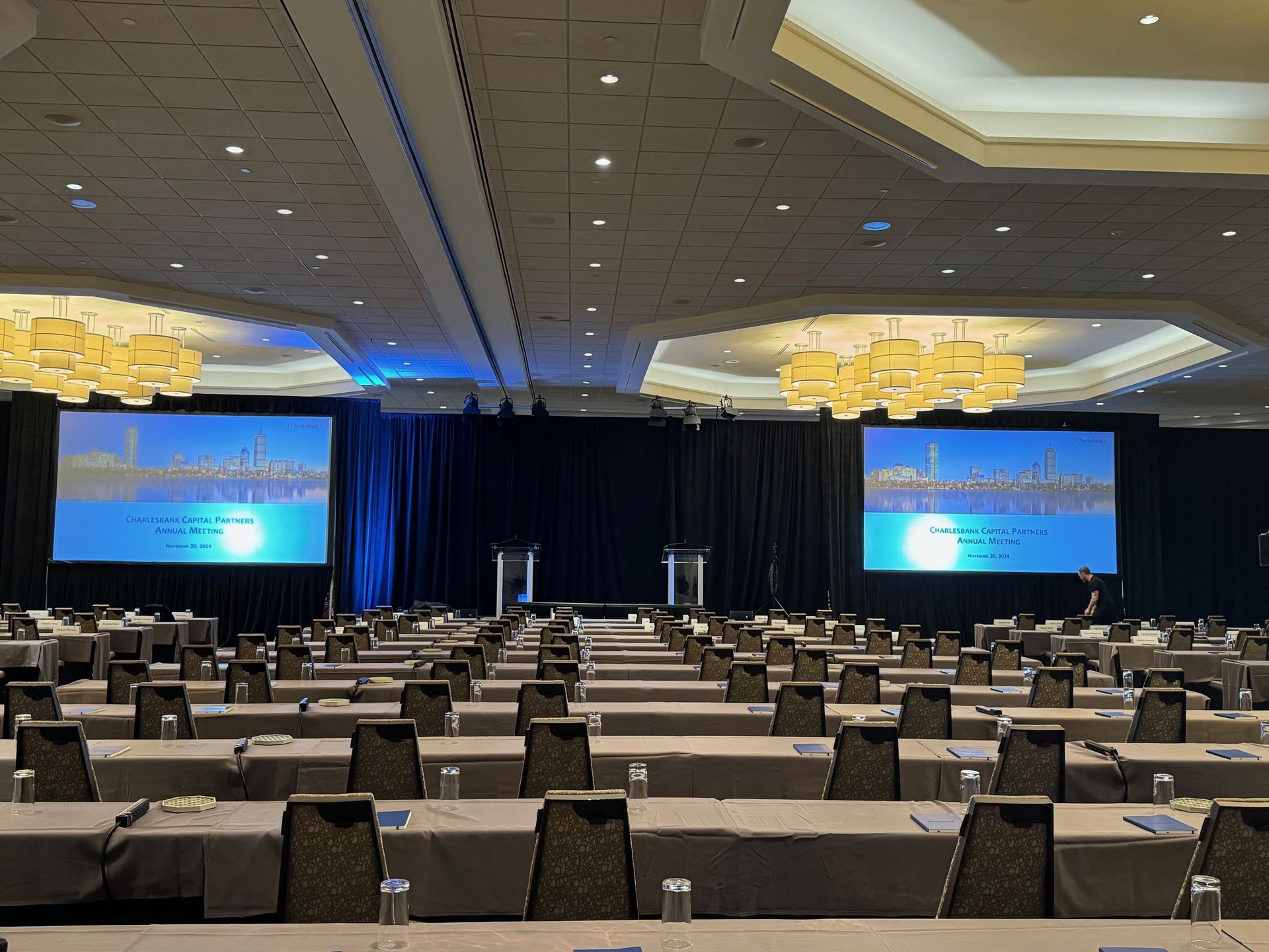 Conference room set up for a presentation. Tables and chairs face a stage with two screens displaying a city skyline.
