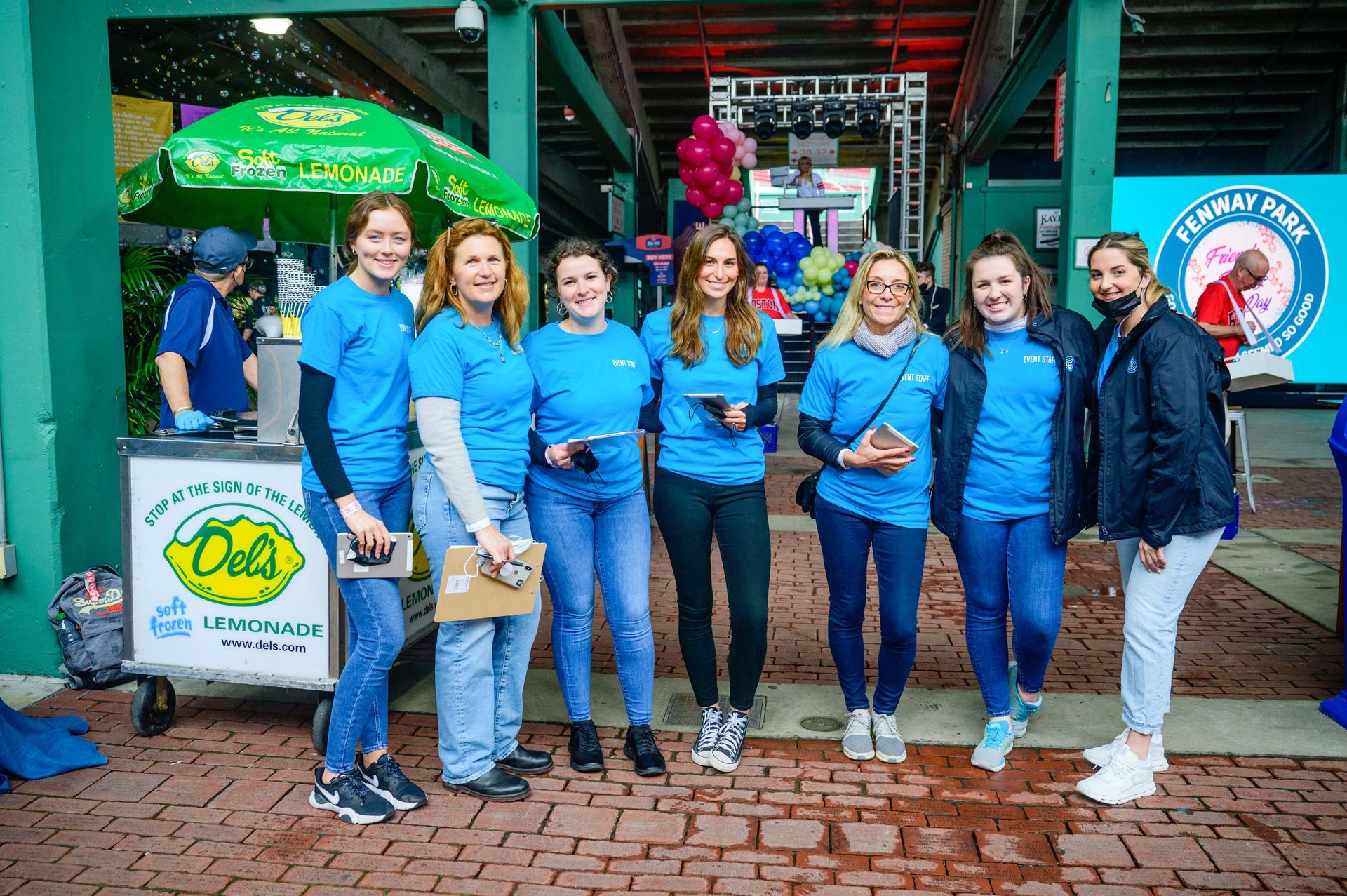 Group of seven people wearing blue shirts, posing in front of a lemonade stand, brick walkway, and a screen.