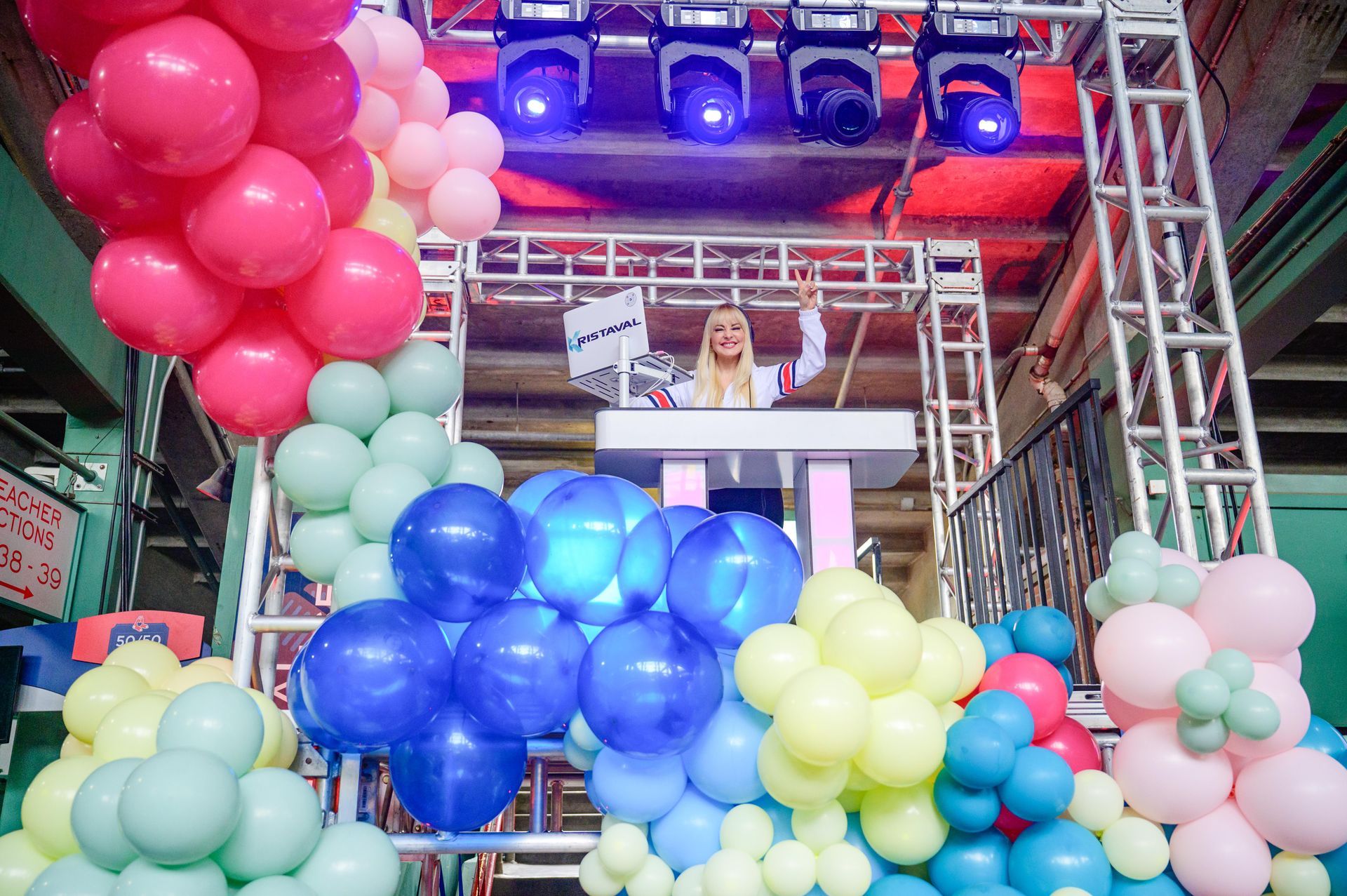 Blonde woman DJing at a party stage with colorful balloons.