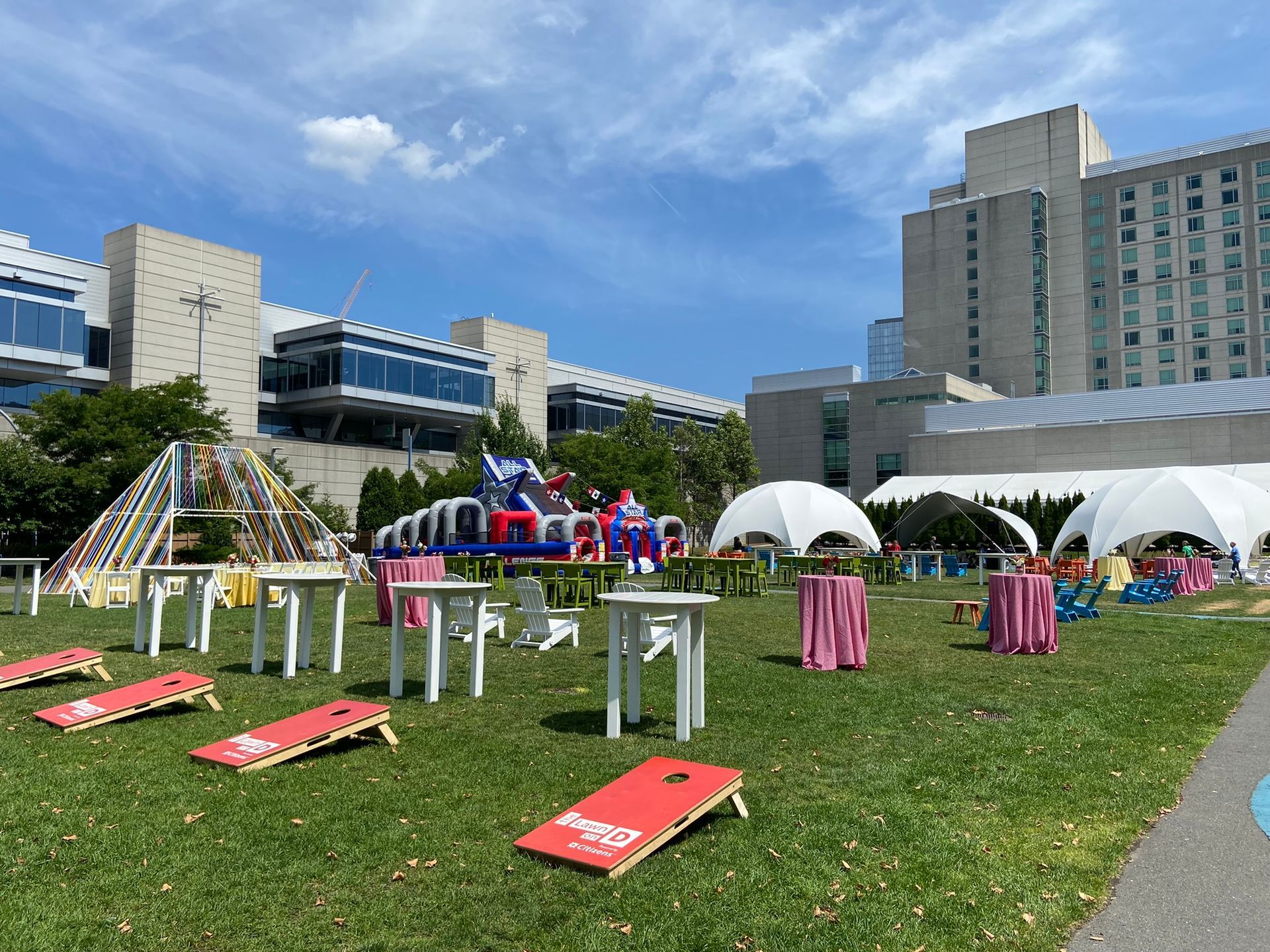 Outdoor event with tables, tents, cornhole, and an inflatable obstacle course on a grassy area in front of buildings.