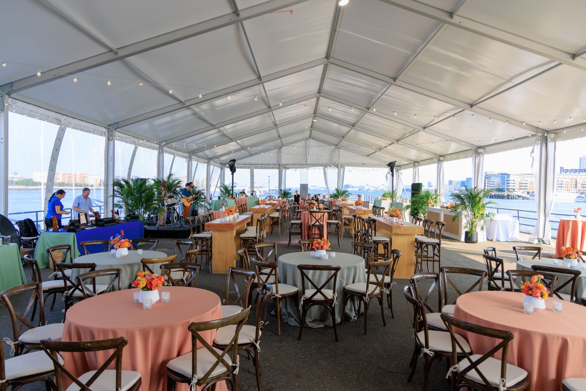 Event tent with round tables and chairs overlooking water; band plays on stage.