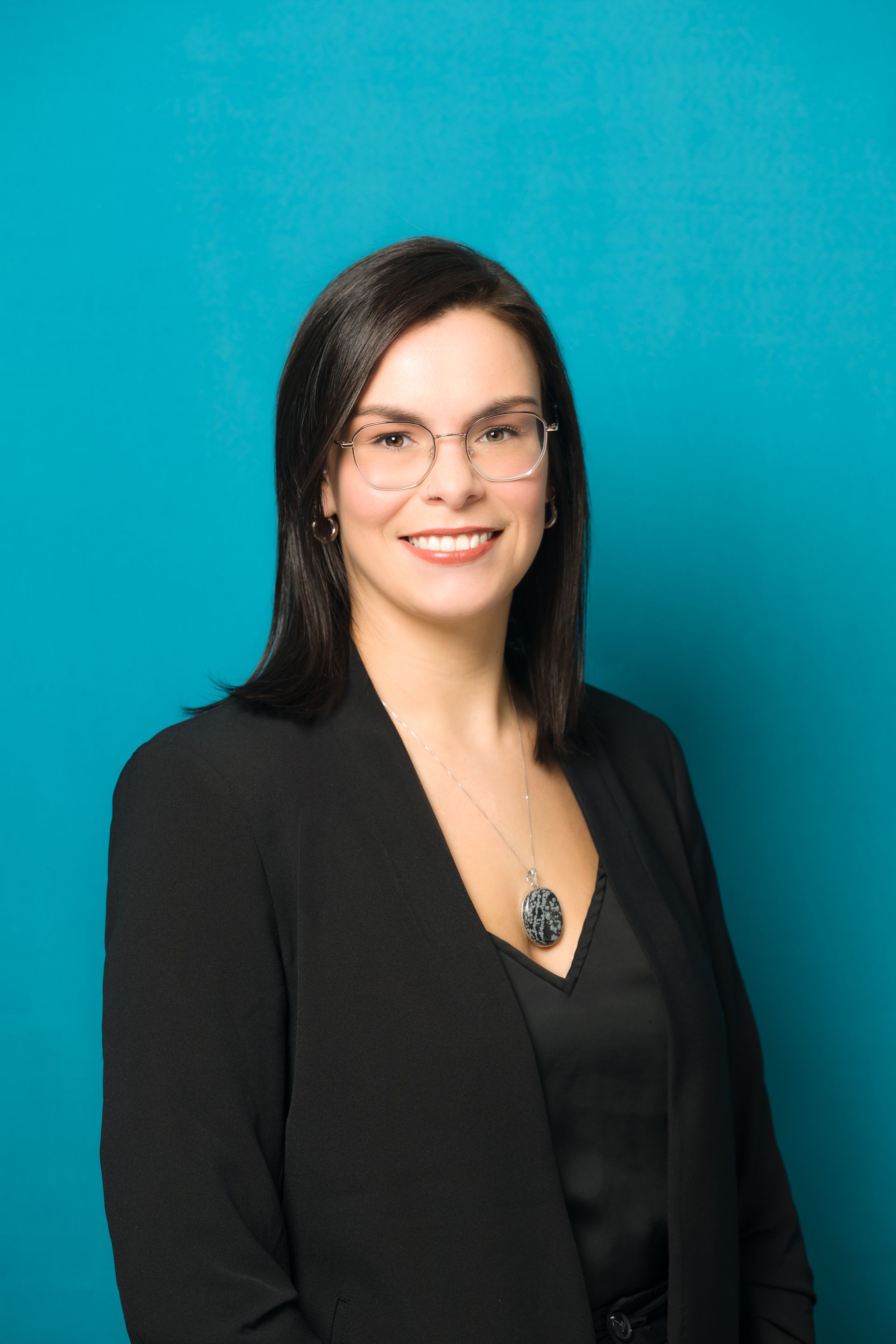 A woman wearing glasses and a black jacket is smiling in front of a blue background.