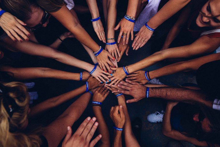 Hands of many people in a circle, touching at center, wearing blue bracelets.