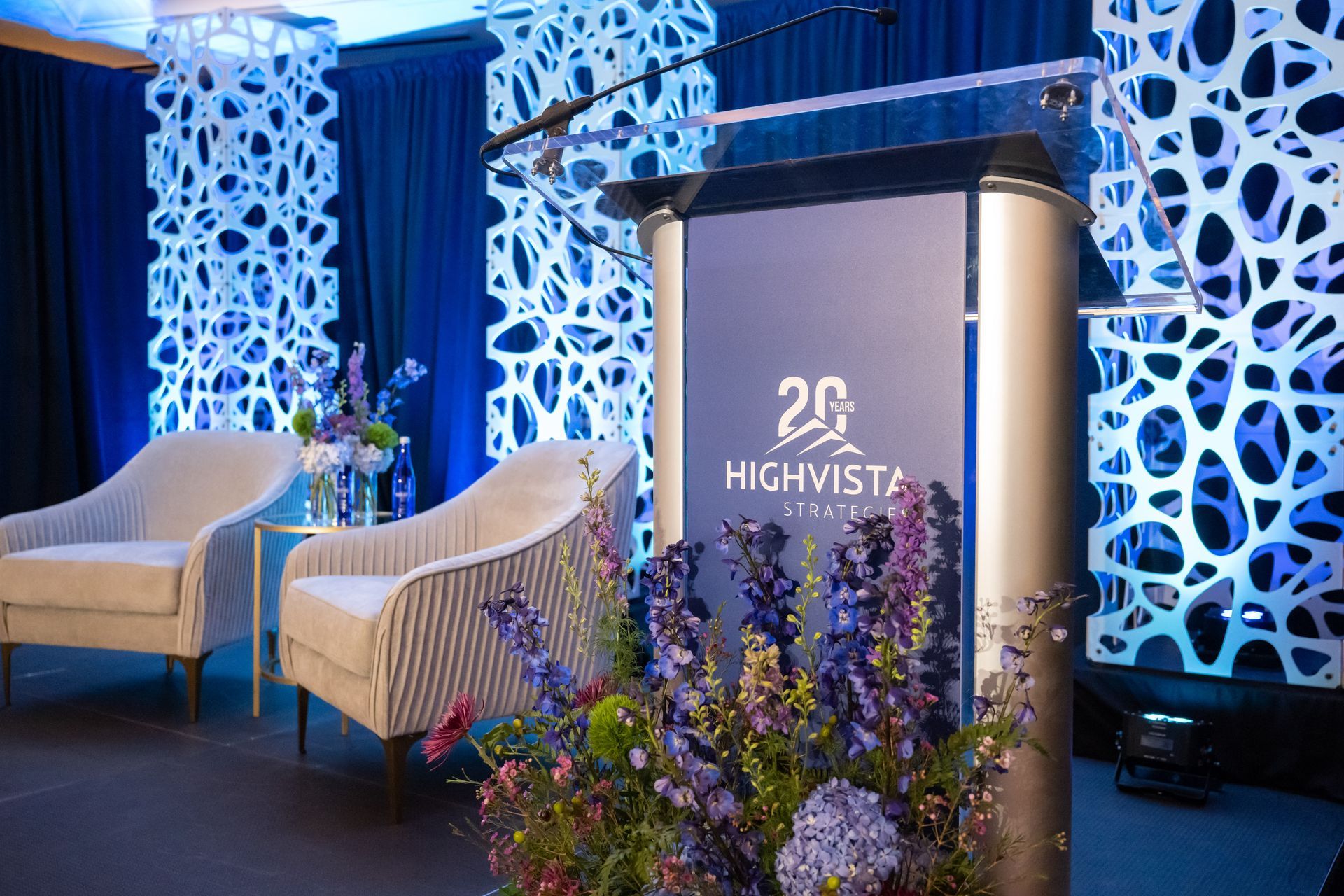 Podium with floral arrangement, two chairs, and decorative blue backdrop for a Highvista event.