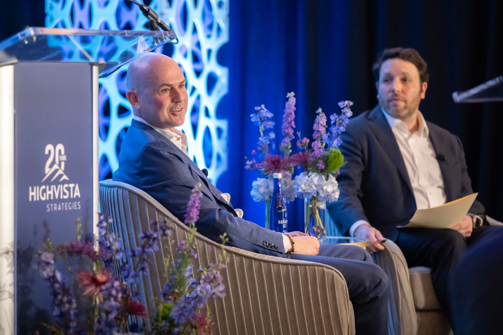 Two men seated on a stage at an event. The man on the left speaks, the other holds papers. Stage backdrop in blue.