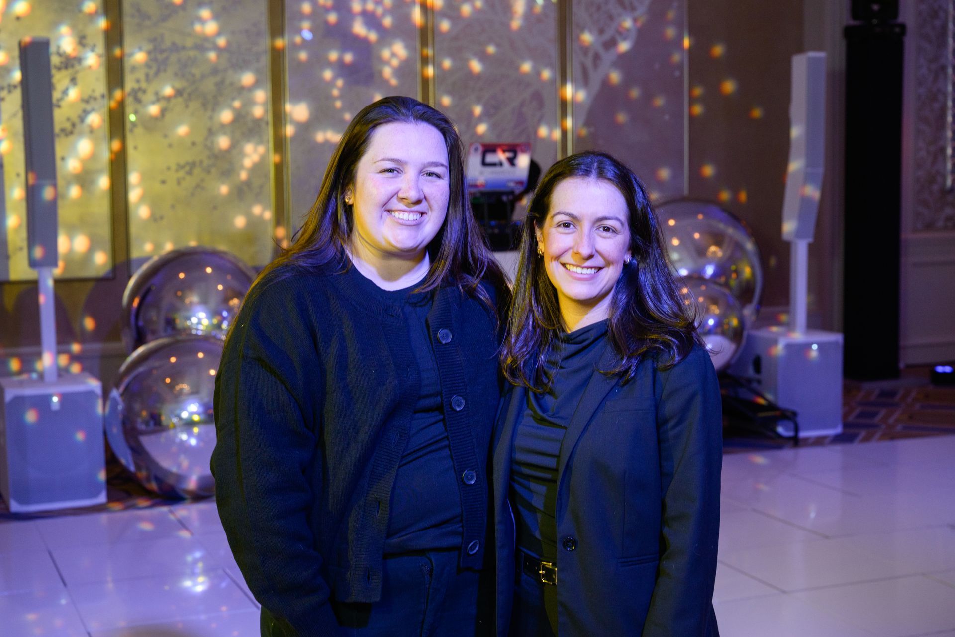 Two women smiling, posing for a photo at an event. Shiny spheres and speakers in background.