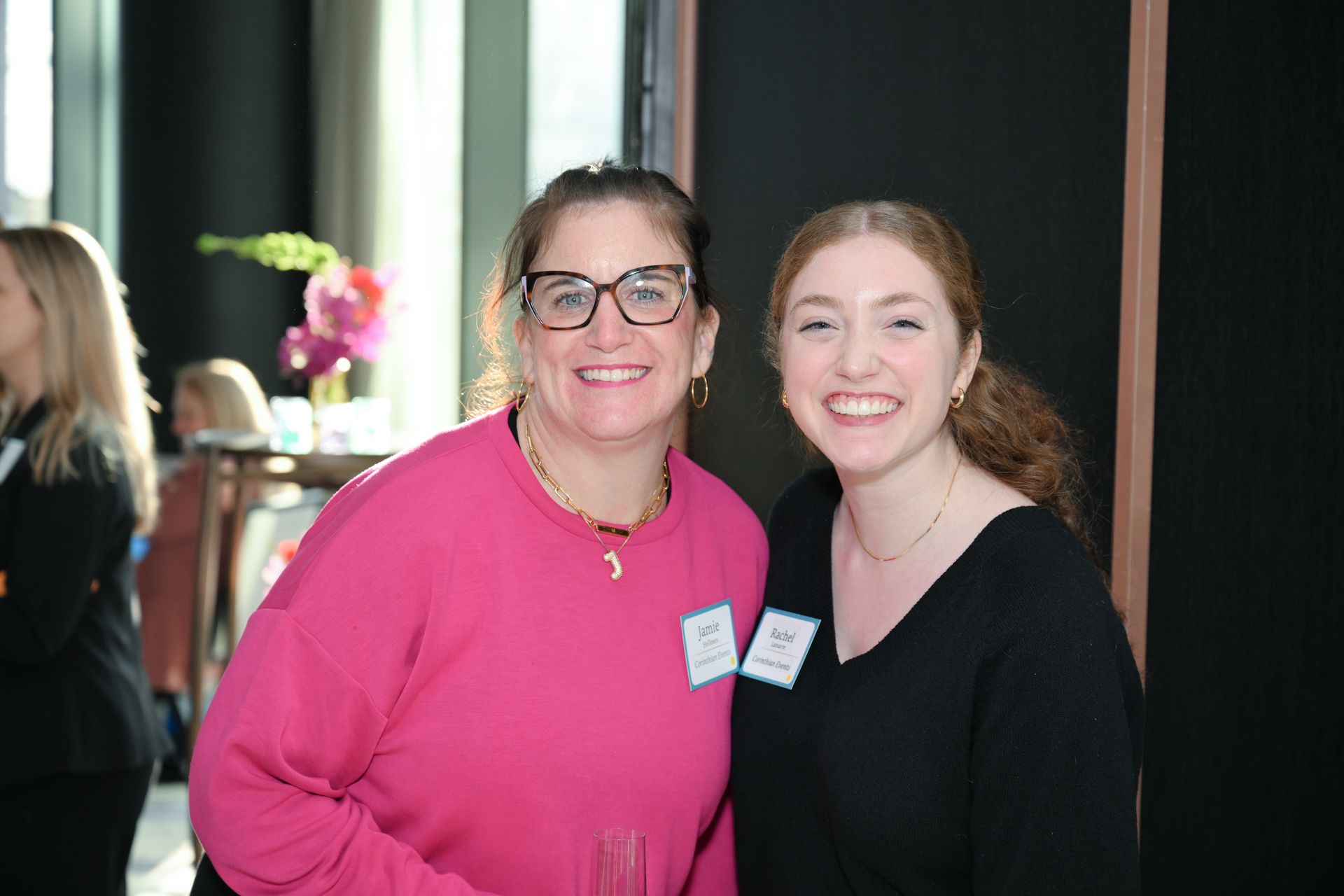 Two women smiling, posing together. One wears a pink sweater and glasses, the other a black top. Indoors, likely an event.