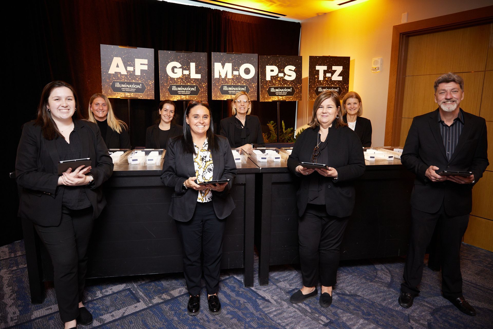 People behind a table with lettered signs, holding tablets, possibly for an event registration.