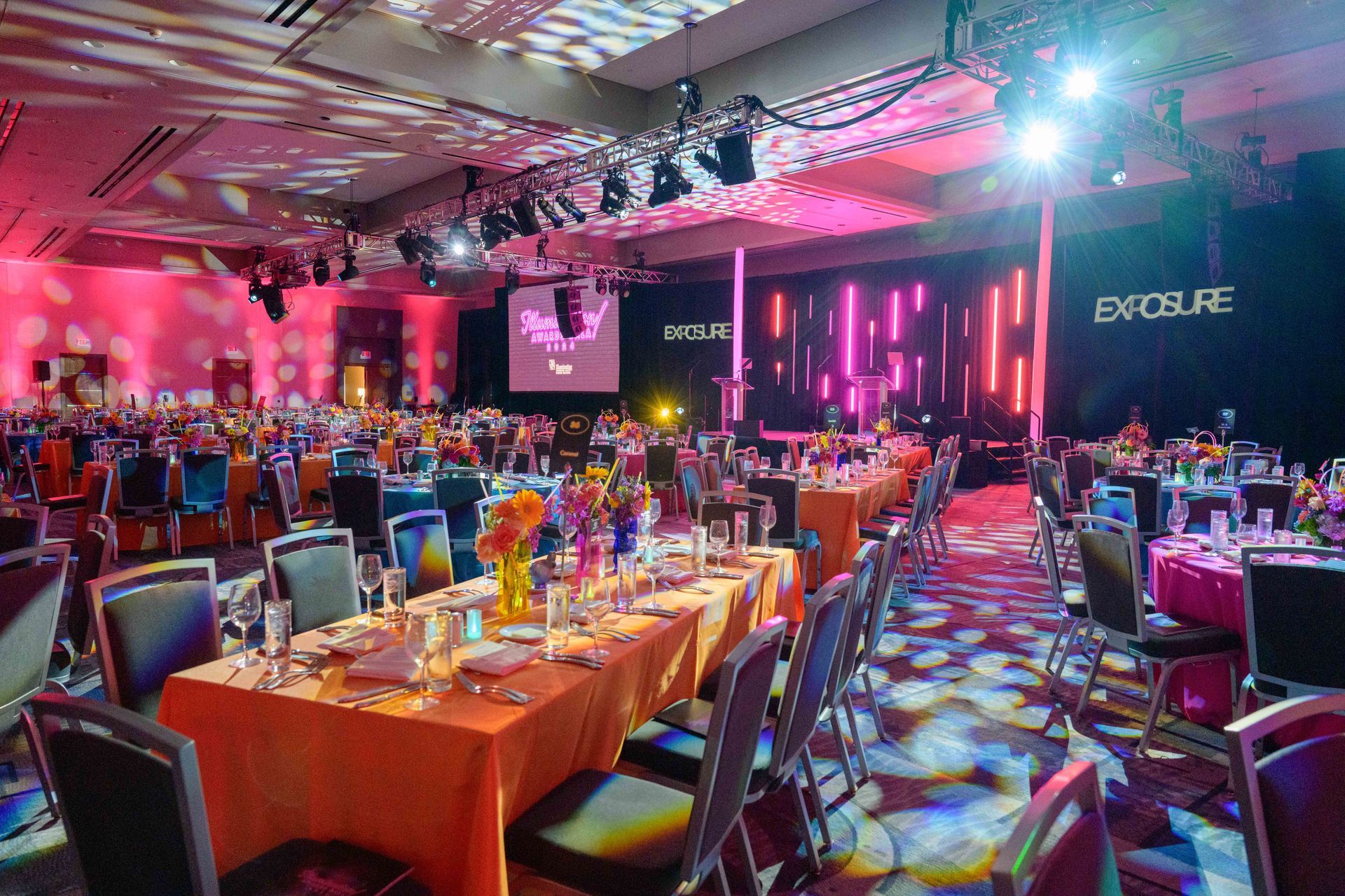 Brightly lit banquet hall set for a gala, featuring decorated tables, stage with "EXPOSURE" logo, and colorful uplighting.