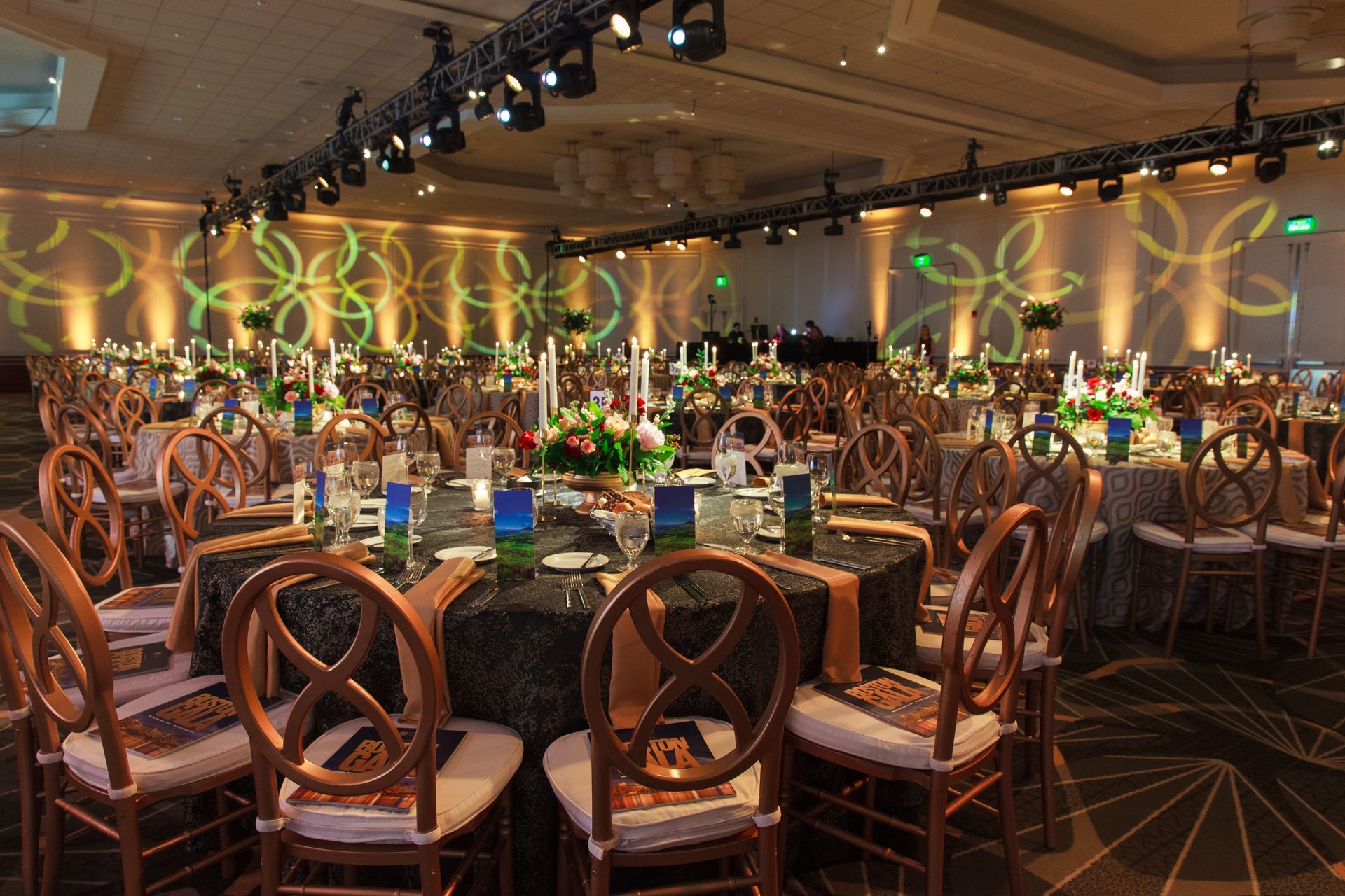 Elegant ballroom set for a formal event. Round tables with gold chairs, floral centerpieces, and patterned tablecloths.