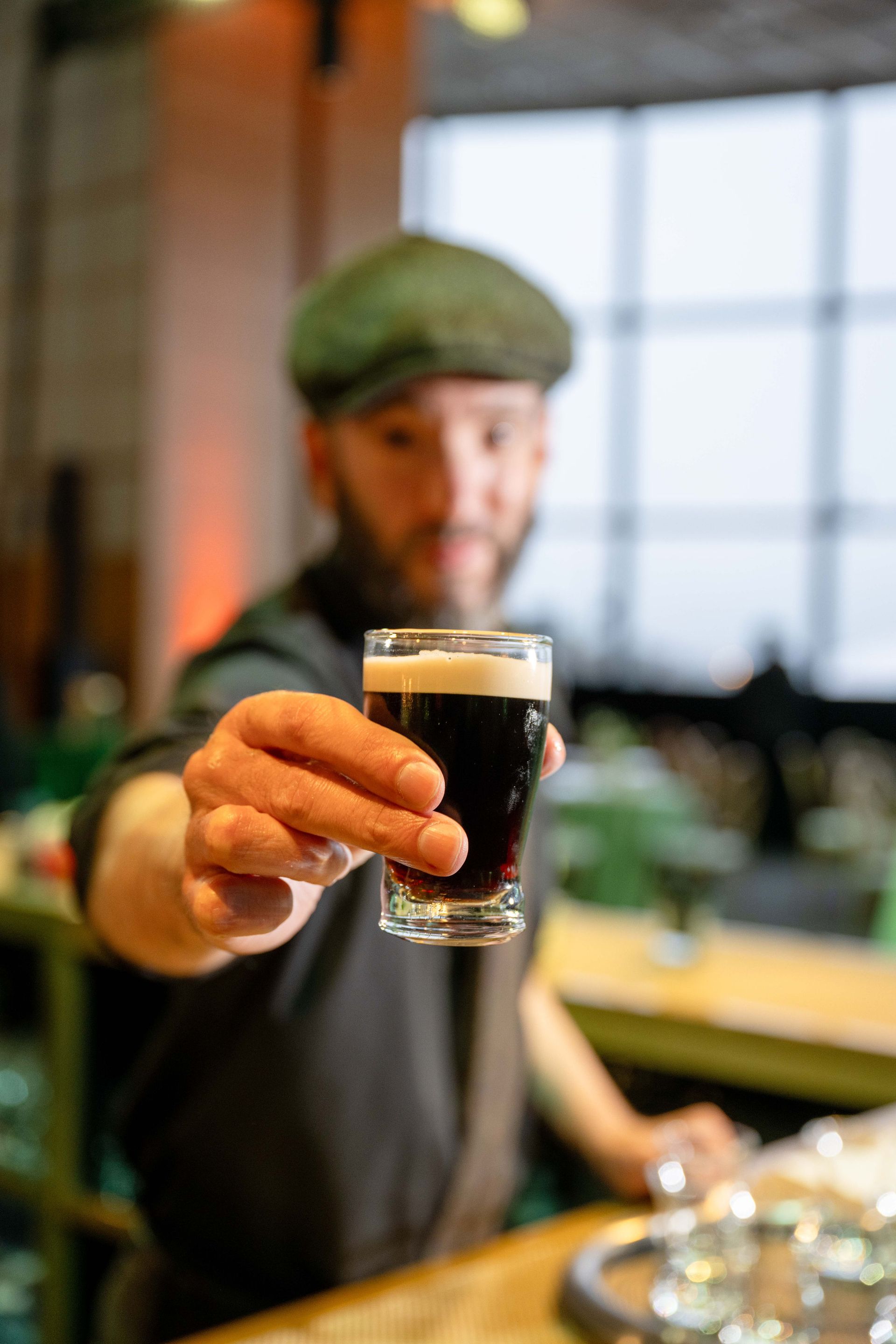 Man in cap holding out a dark beer, interior shot with blurry background.