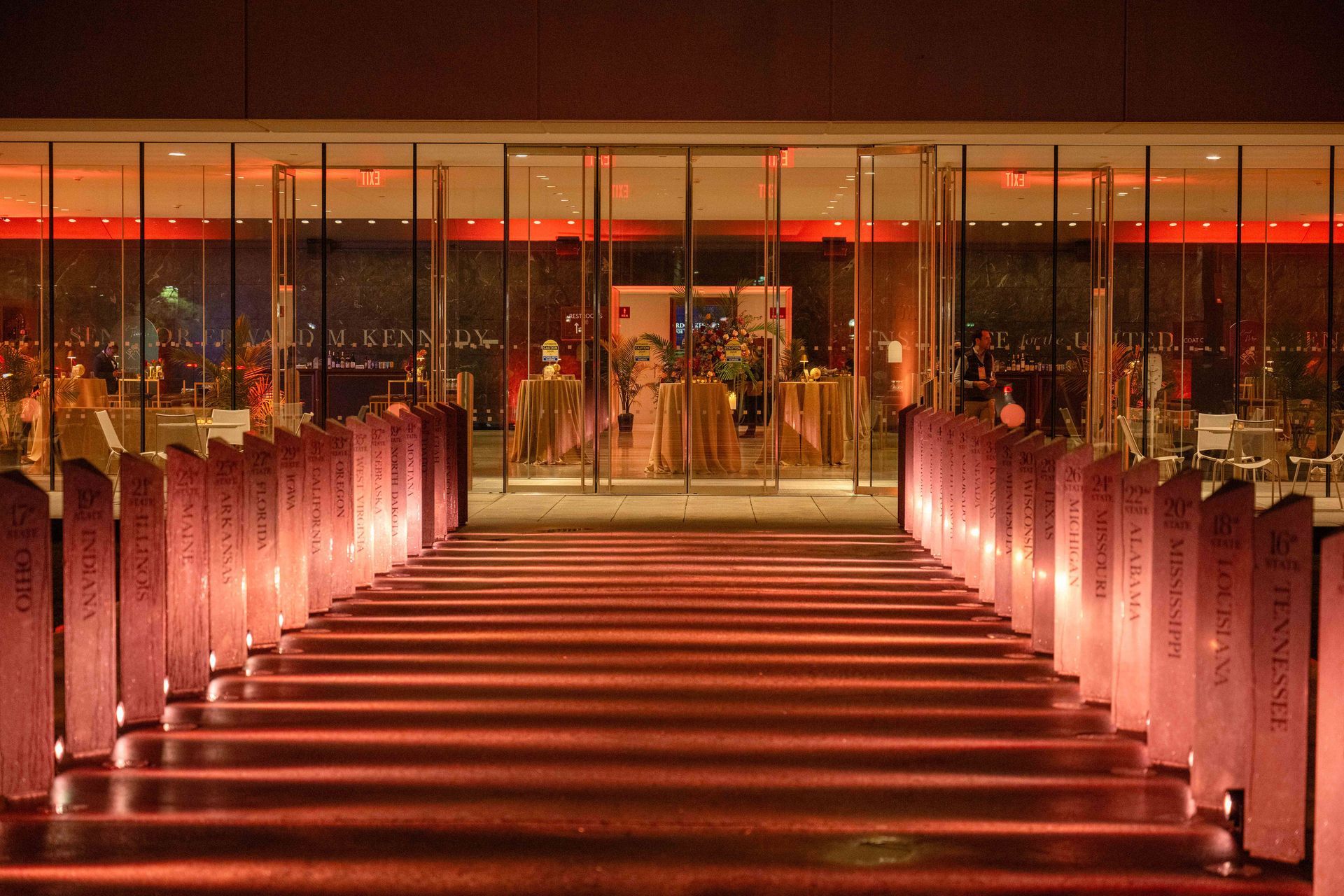 Stairway leading to a glass building, illuminated by red lights, with columns on both sides.