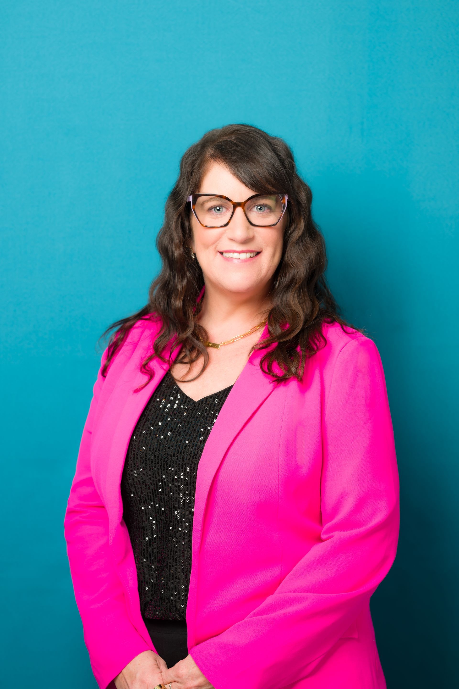 Woman in glasses, pink blazer, black top, smiling against a teal background.