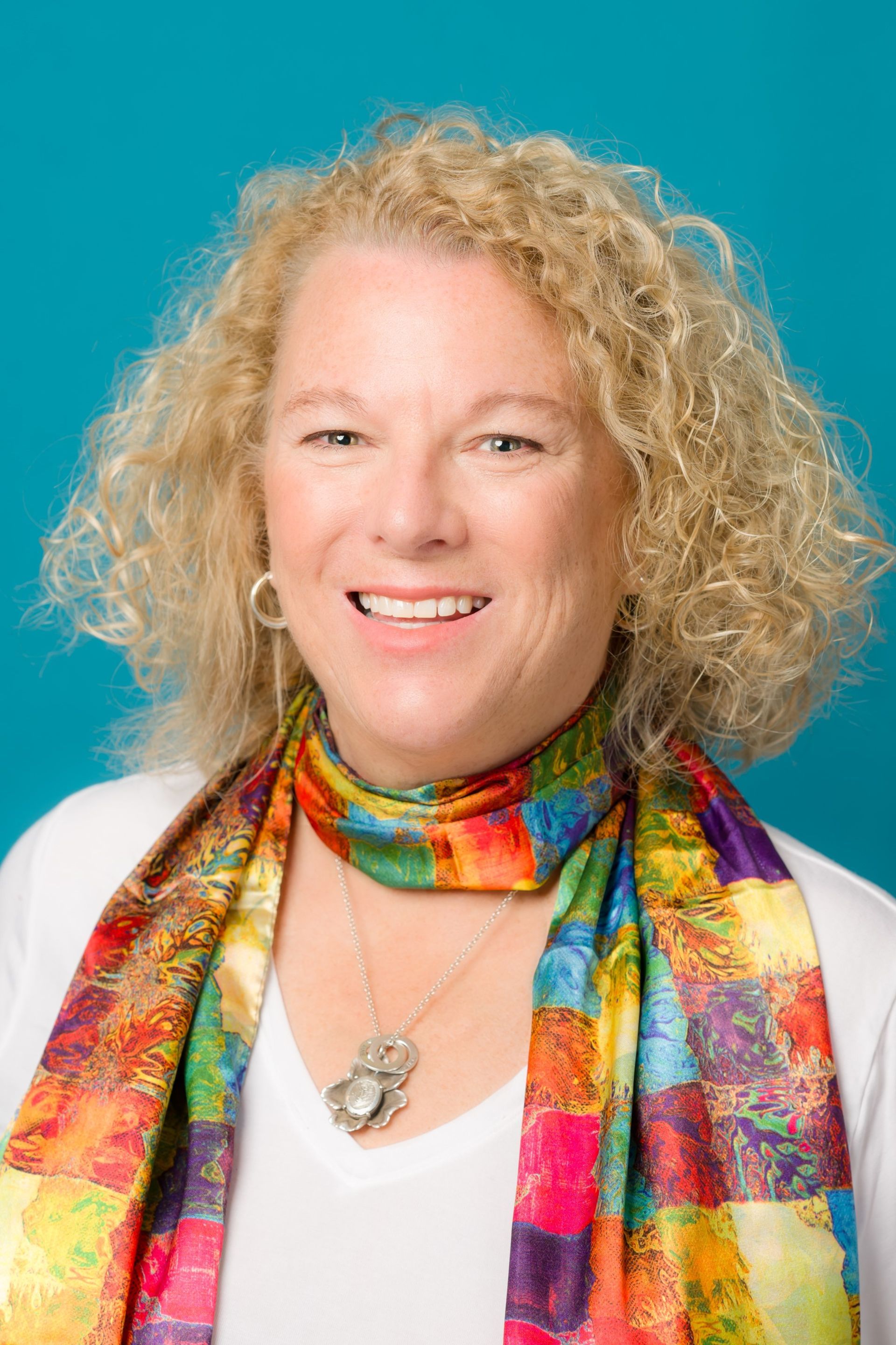 Woman with blonde curly hair smiles, wearing a colorful scarf and pendant, against a teal background.