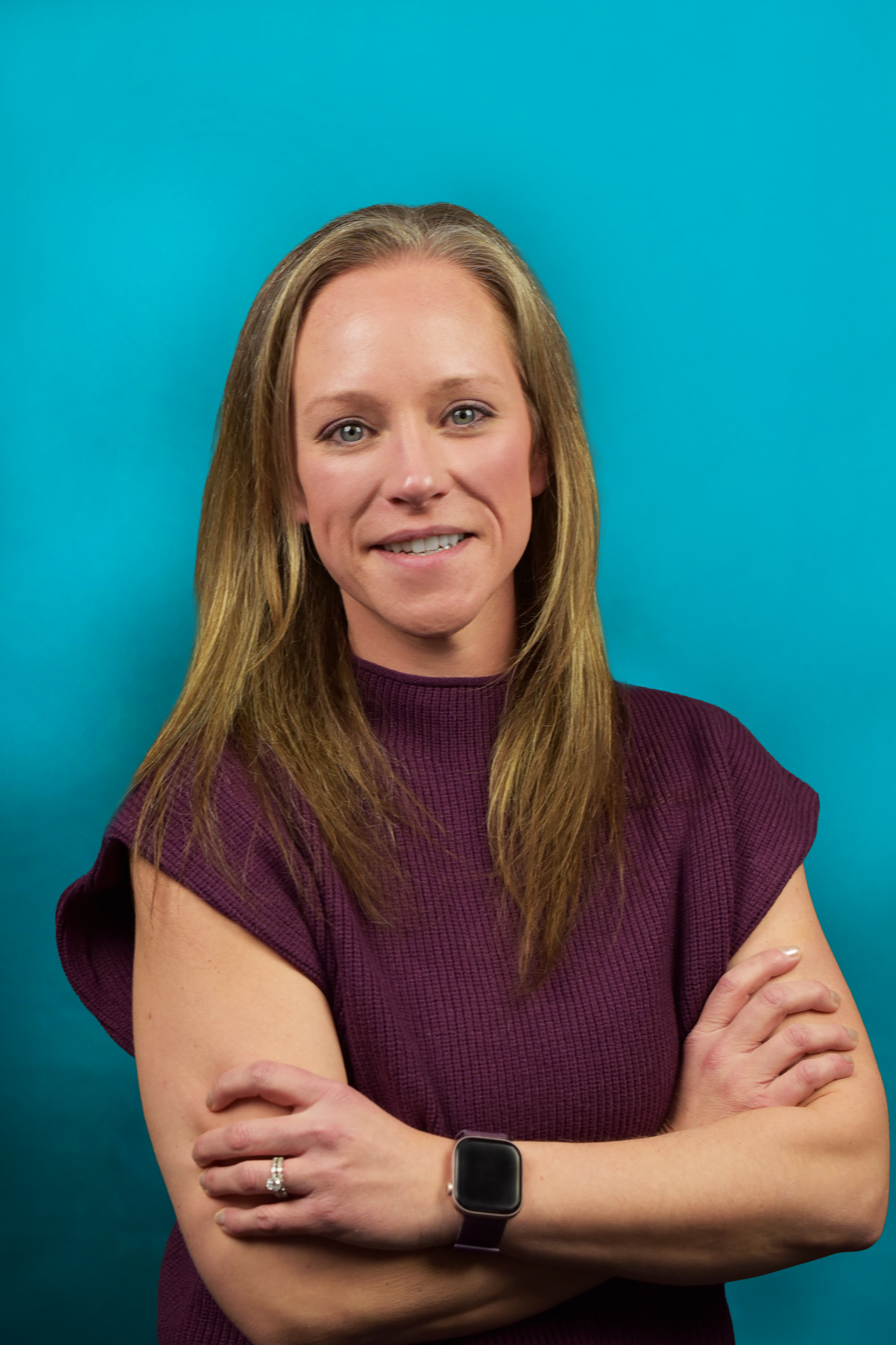 Woman in purple sweater with arms crossed, smiling, against a teal background.