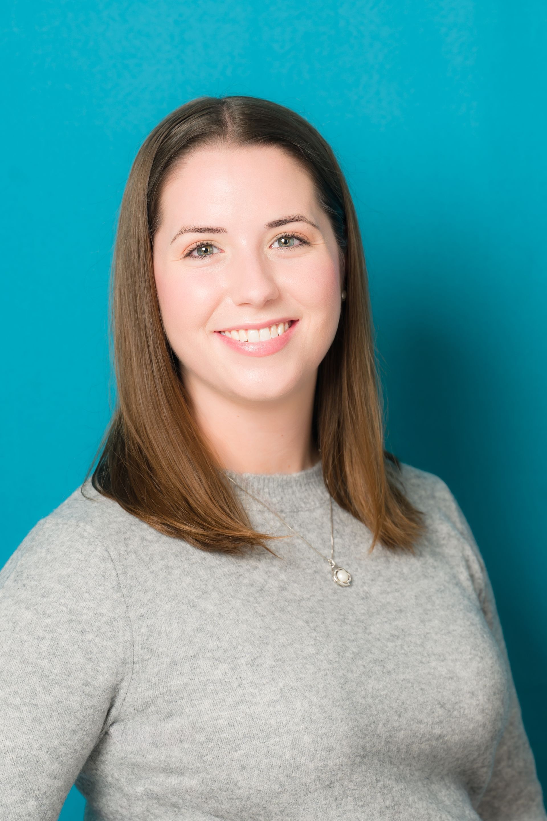 A woman in a grey sweater is smiling in front of a blue background.