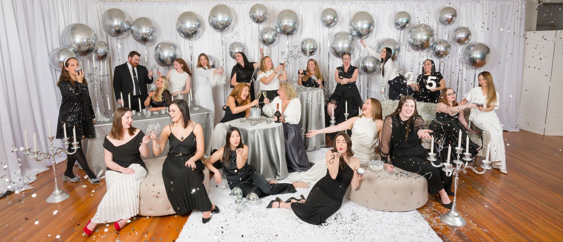 Group of people celebrating with silver balloons and confetti; some are drinking and posing.