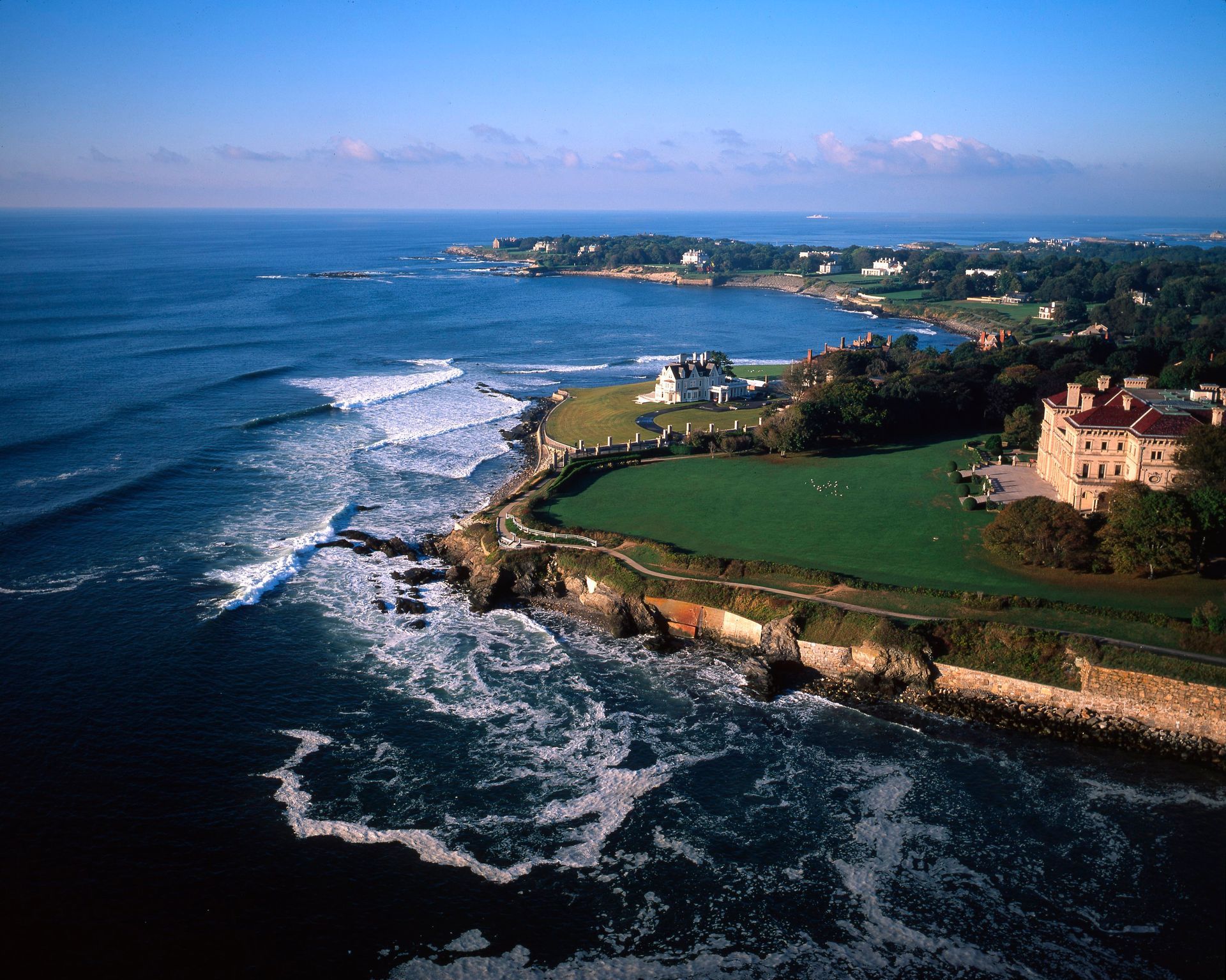 Coastal view of Newport, Rhode Island, with ocean waves crashing against a rocky shore and mansions on a green lawn.