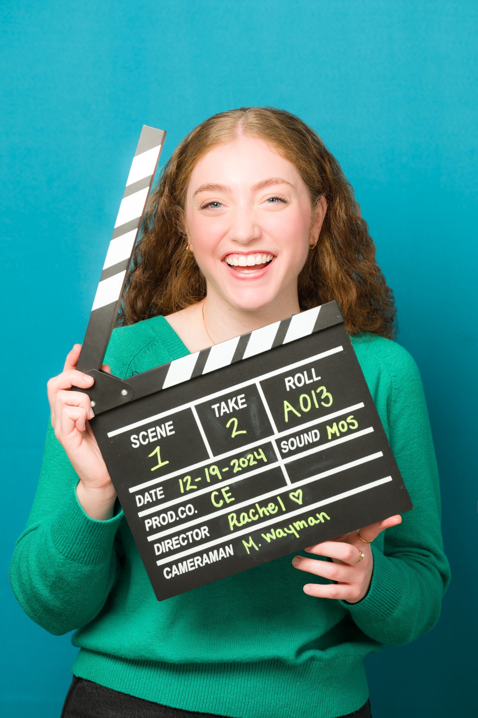 A young woman in a green sweater is holding a clapper board.