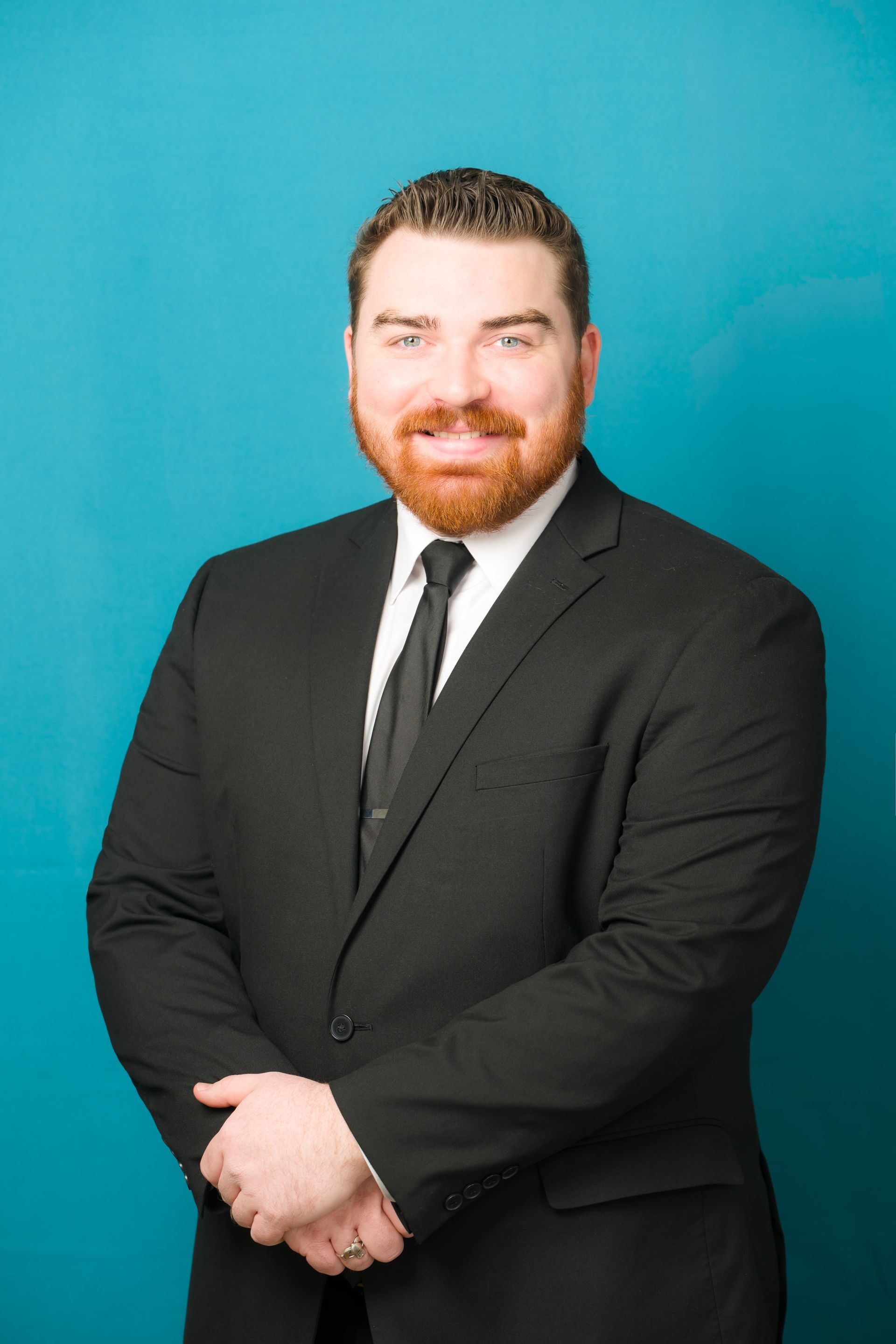 Man in black suit and tie, hands clasped, smiling against a teal background.