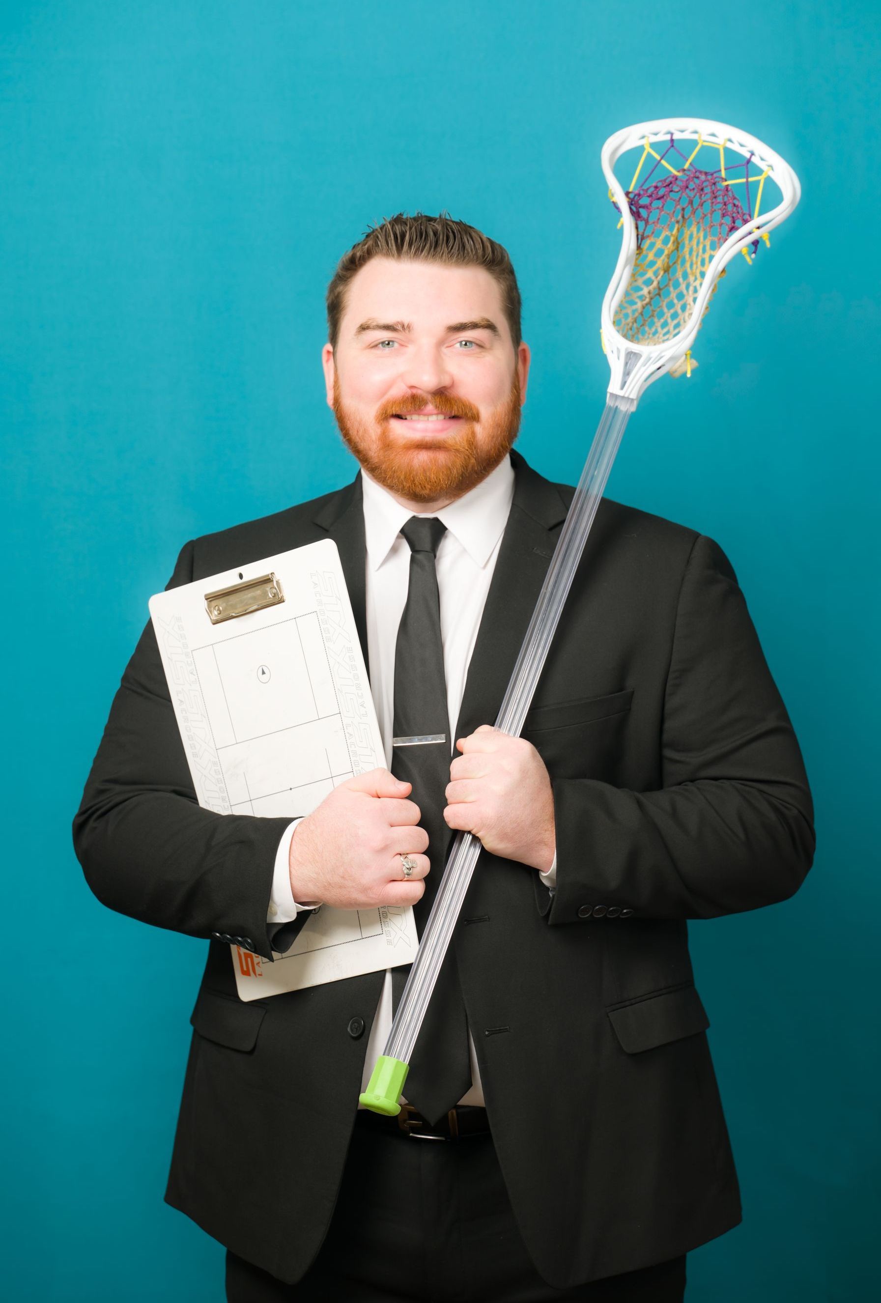 Man in a suit holding a clipboard and lacrosse stick against a teal background.