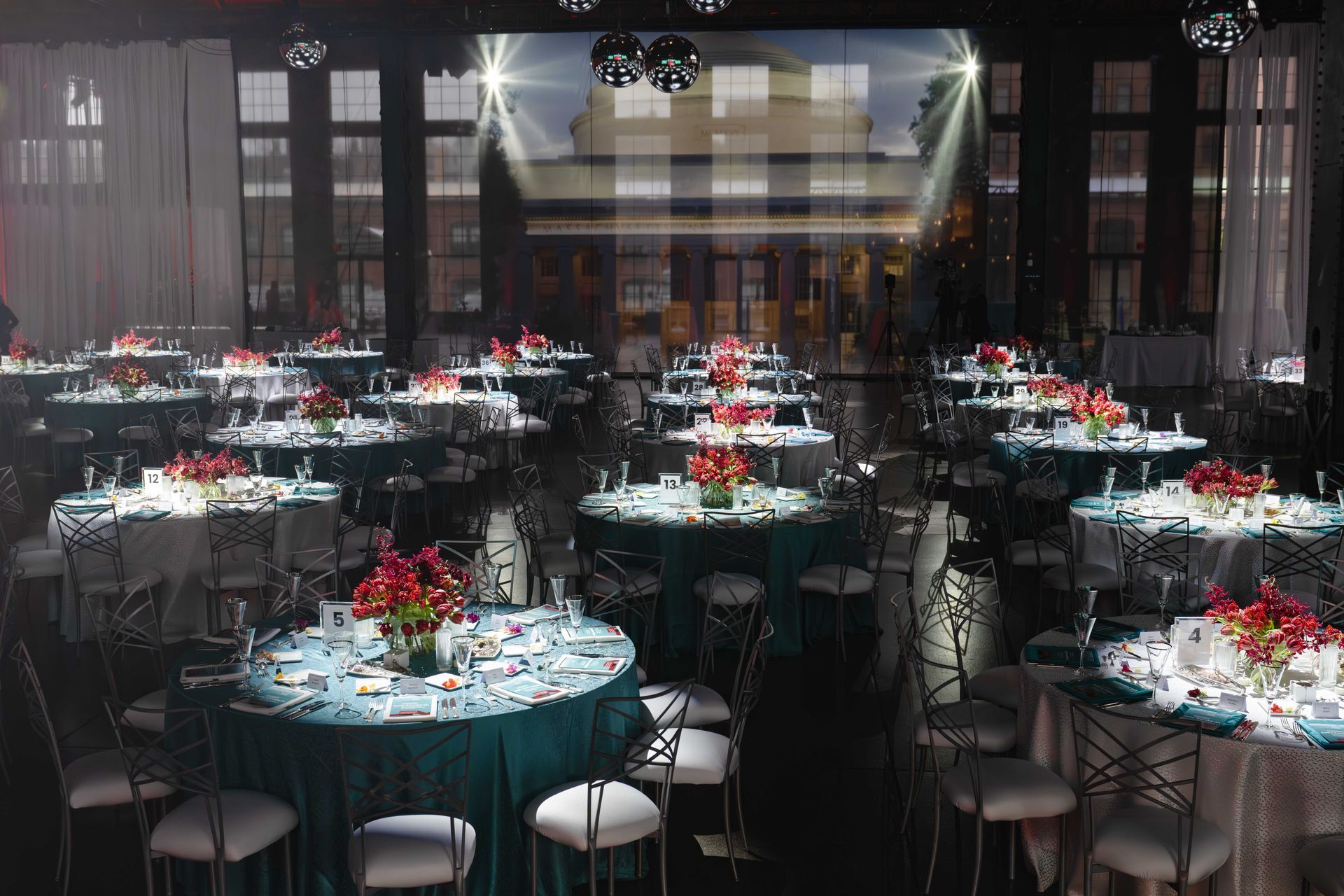 Formal event hall with round tables, teal and white linens, red floral centerpieces, and mirrored floor.