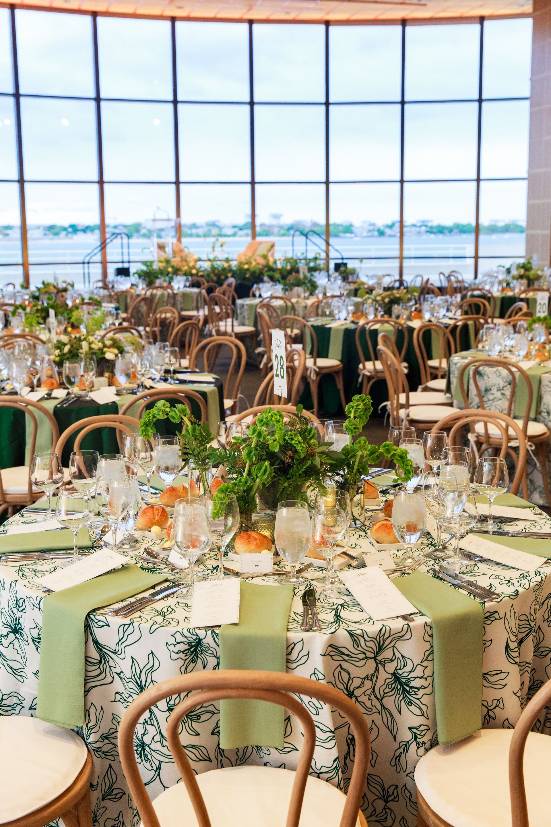 Formal event tables set in a room with large windows, floral tablecloths, green napkins, and decor, overlooking water.