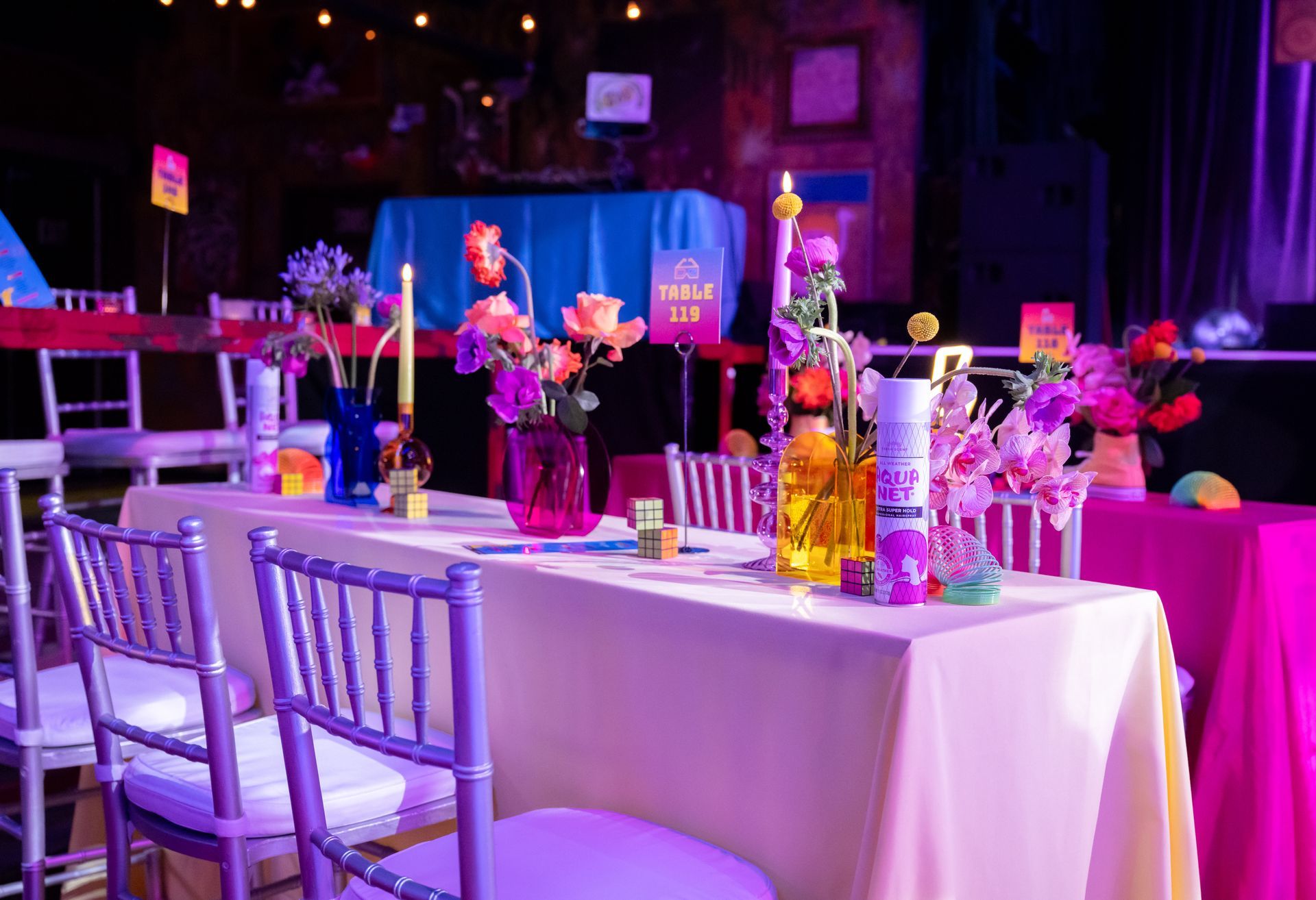 Decorated tables with colorful flowers, candles, and yellow tablecloths, set for an event with purple lighting.