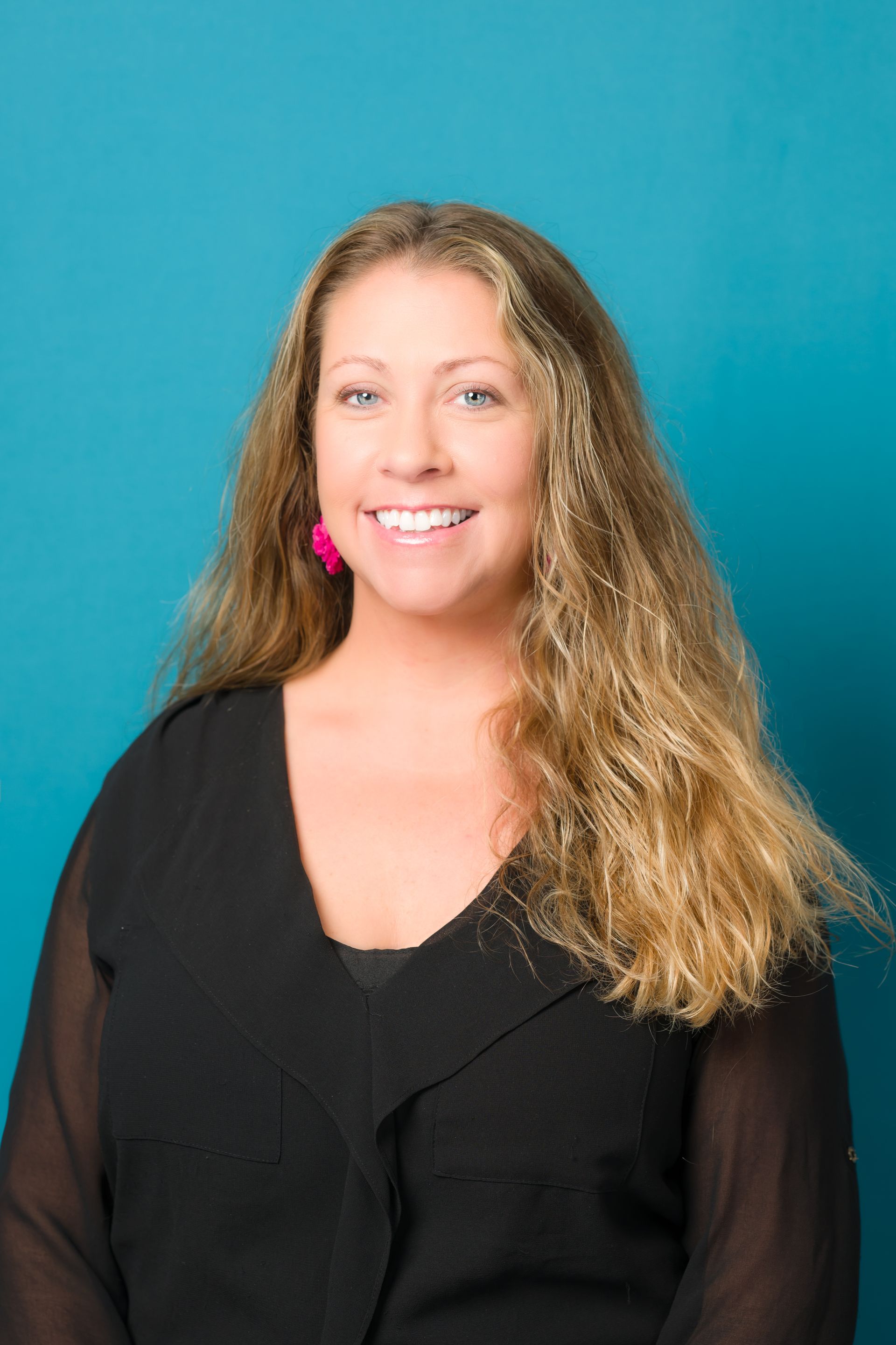 Woman smiling, blonde hair, wearing a black top, pink earrings; against a blue background.