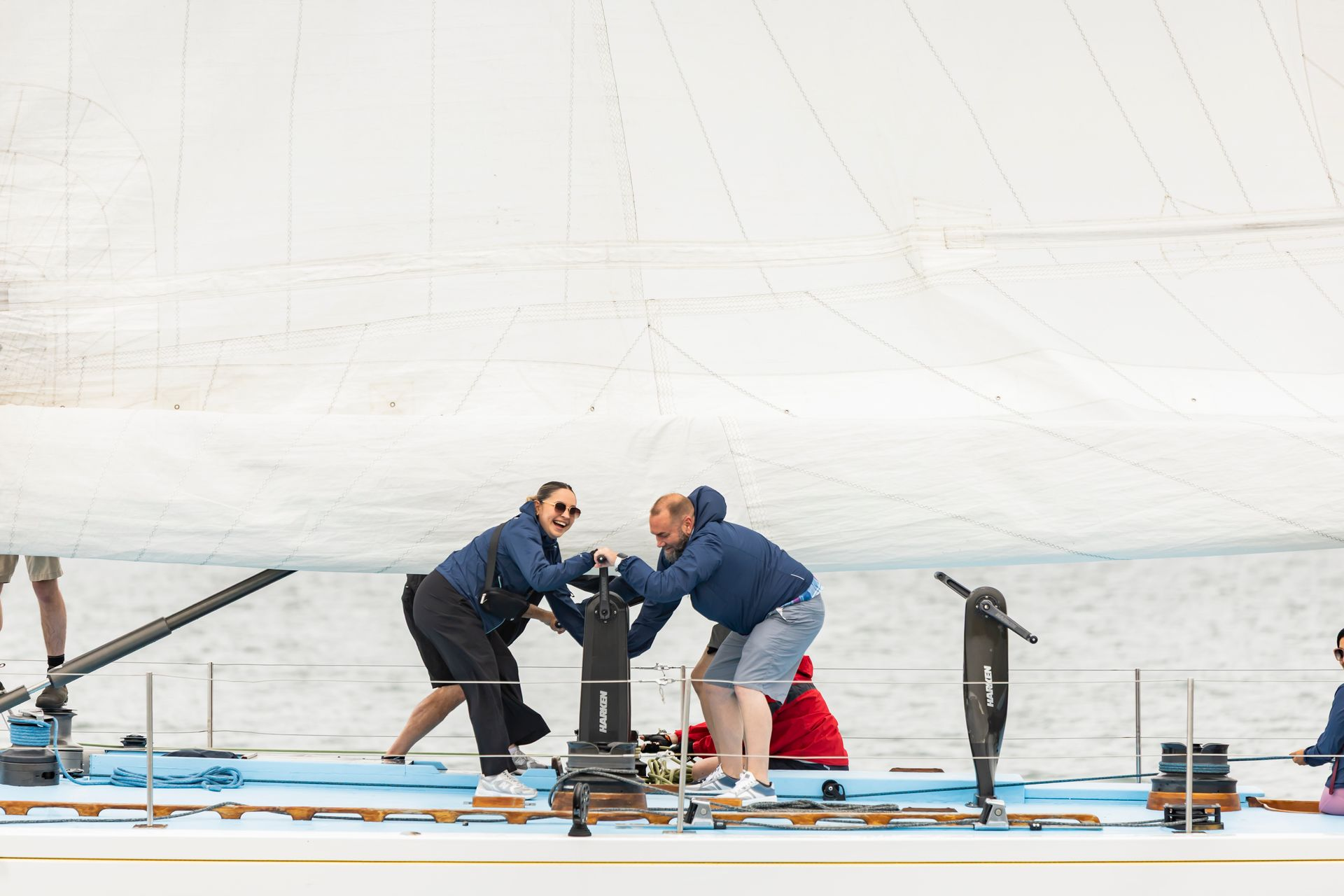 Two people pulling on a yacht's winch. They are on a boat with a white sail visible in the background.