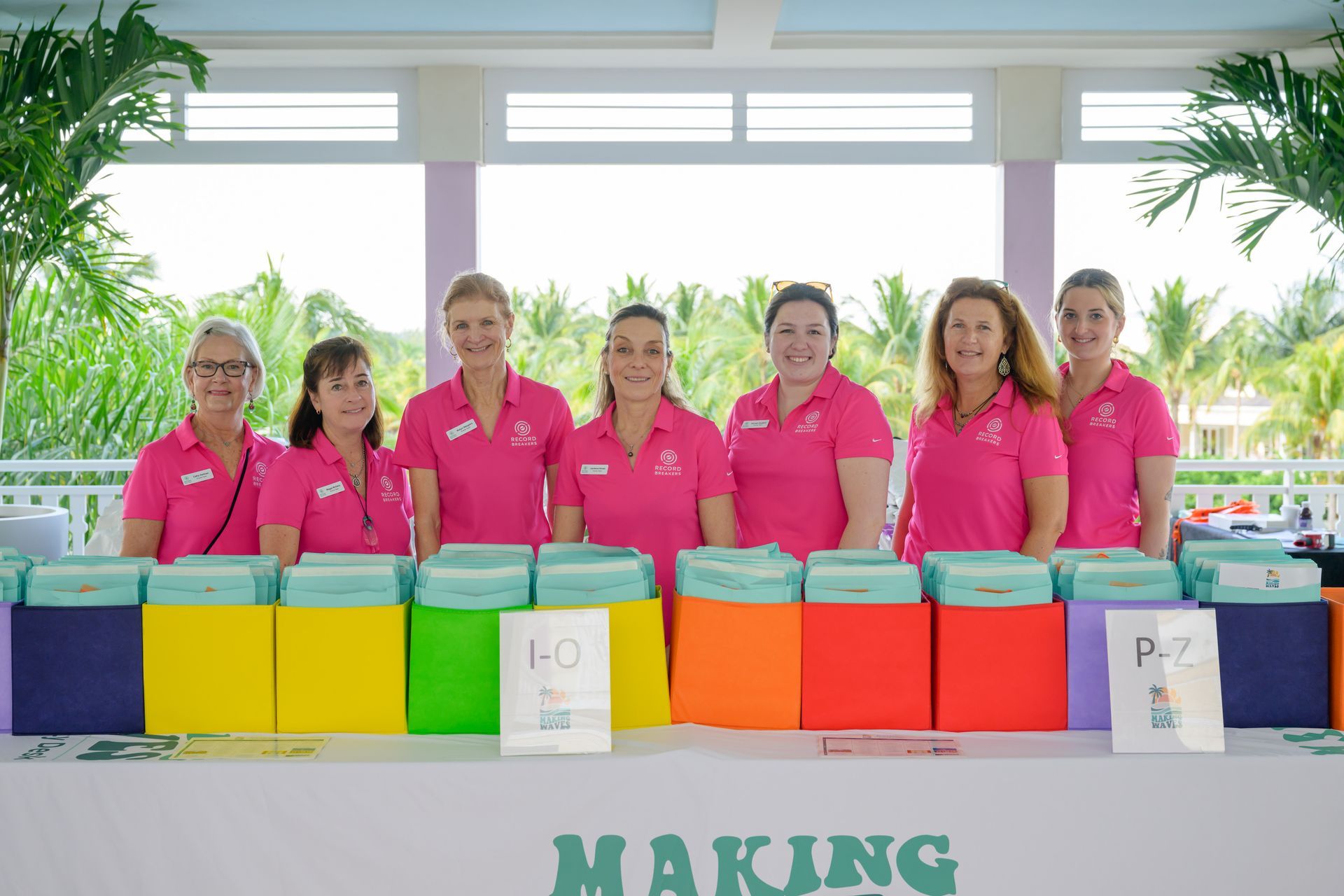 Seven women in pink shirts behind a table with colorful boxes and stacks of blue items.
