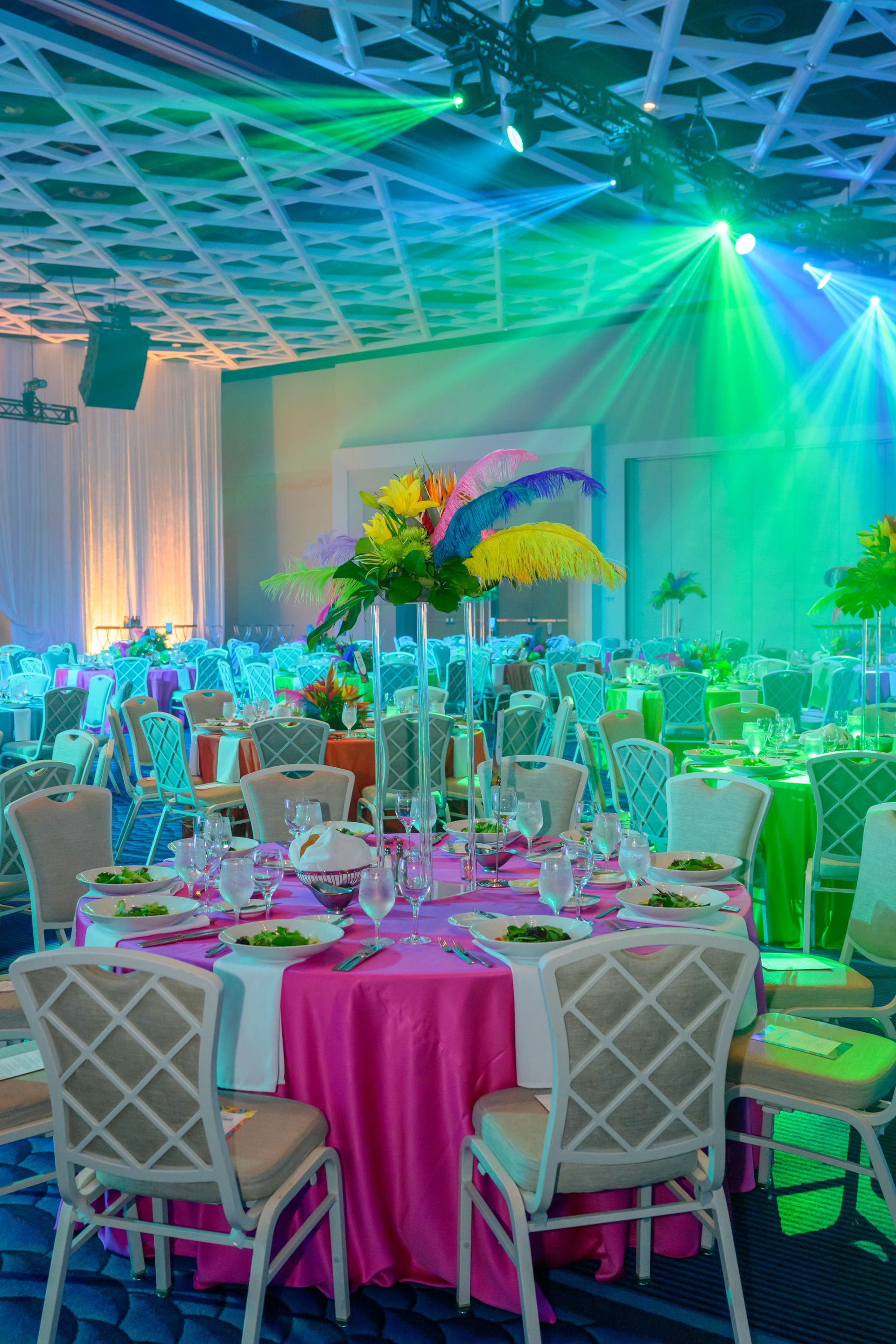 Formal event with tables set, vibrant pink tablecloths, and colorful floral centerpieces.  Green and blue lighting.