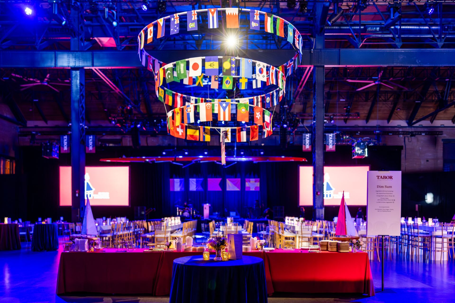 Banquet hall with tables, international flags hanging, stage, and red/blue lighting.