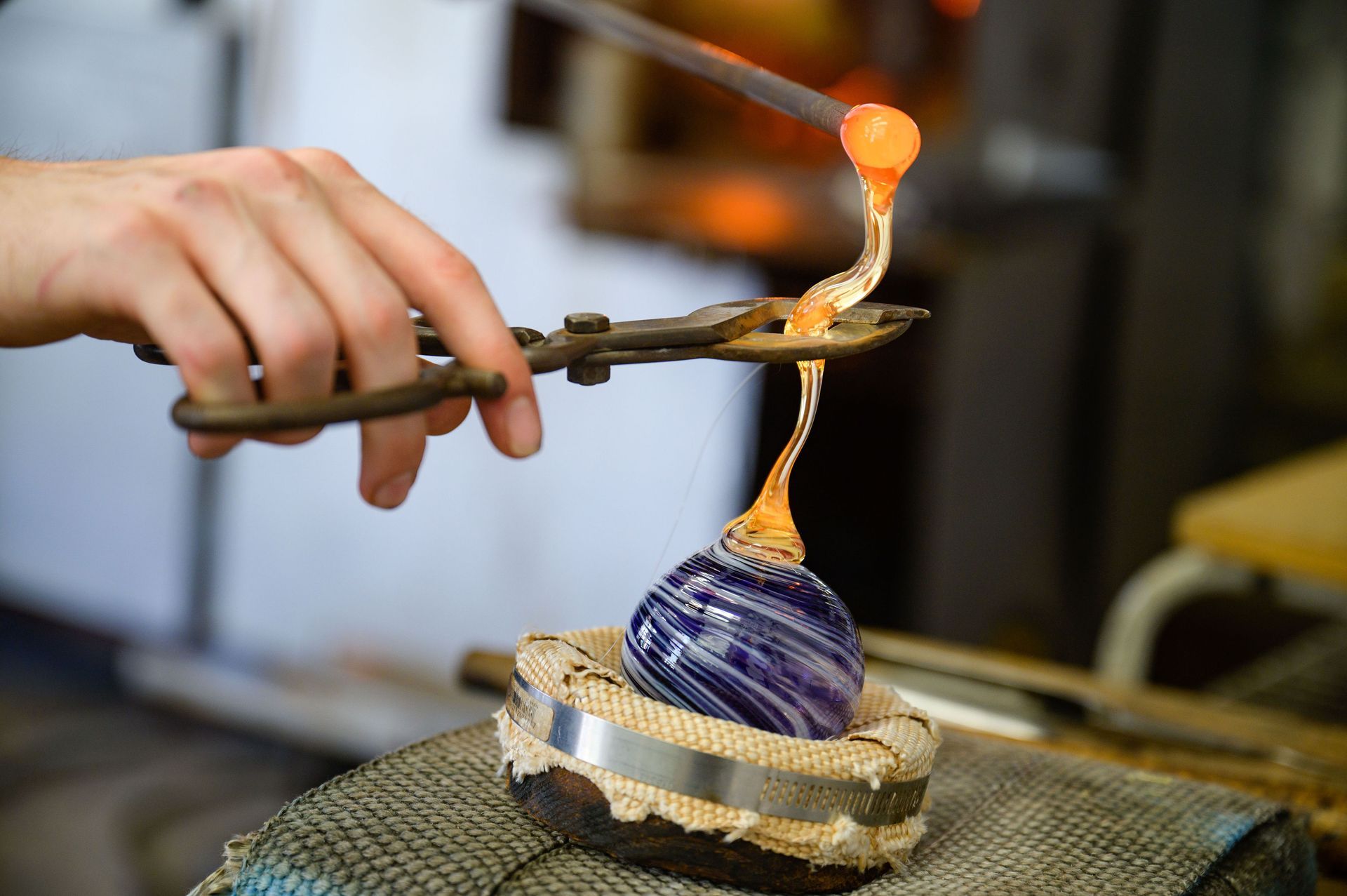 Glassblower using tongs to shape molten glass onto a blue and purple swirled glass form.