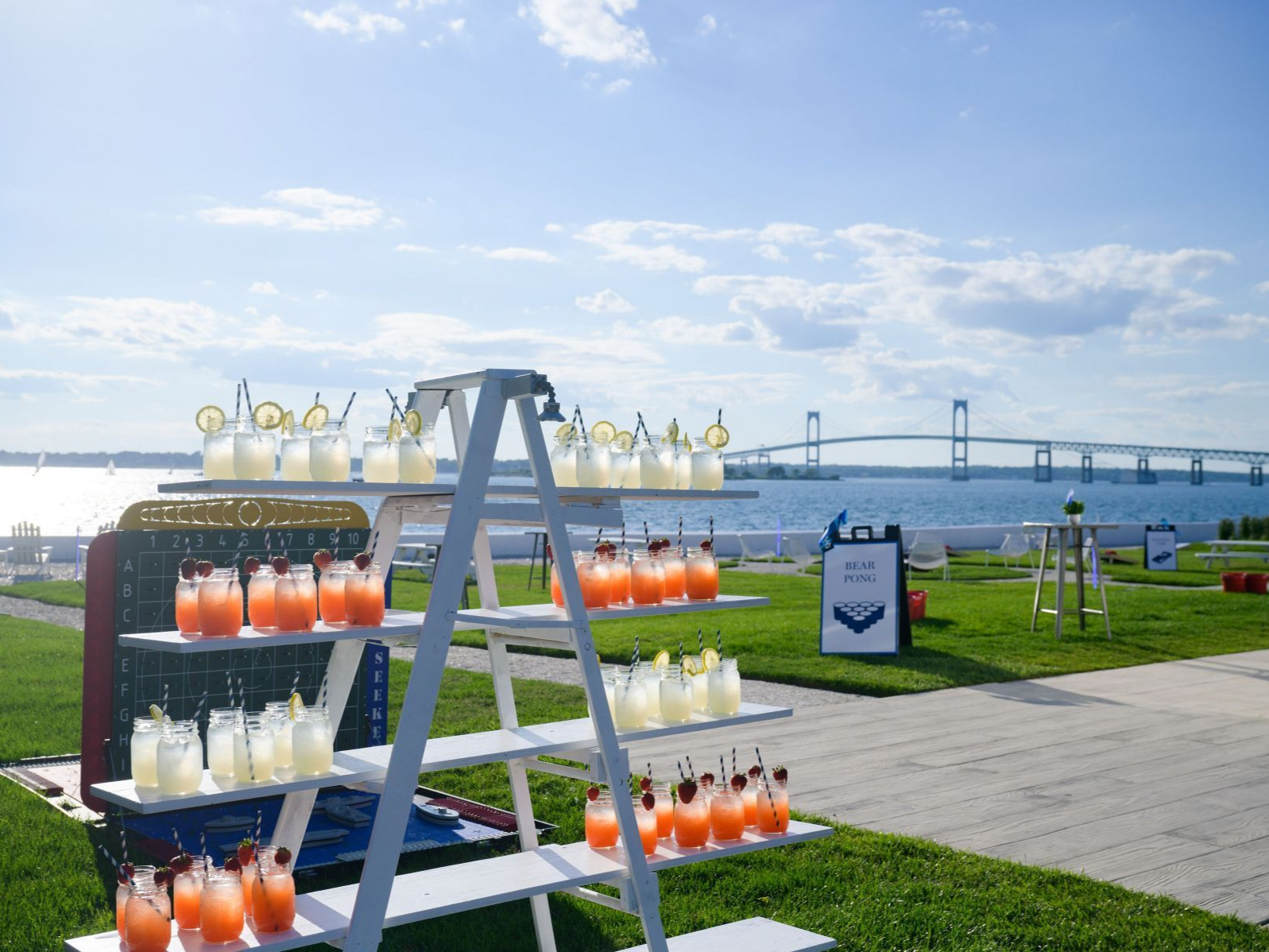 Cocktail bar with tiered shelves outdoors, overlooking a bridge and water. Drinks are various colors, sunny day.