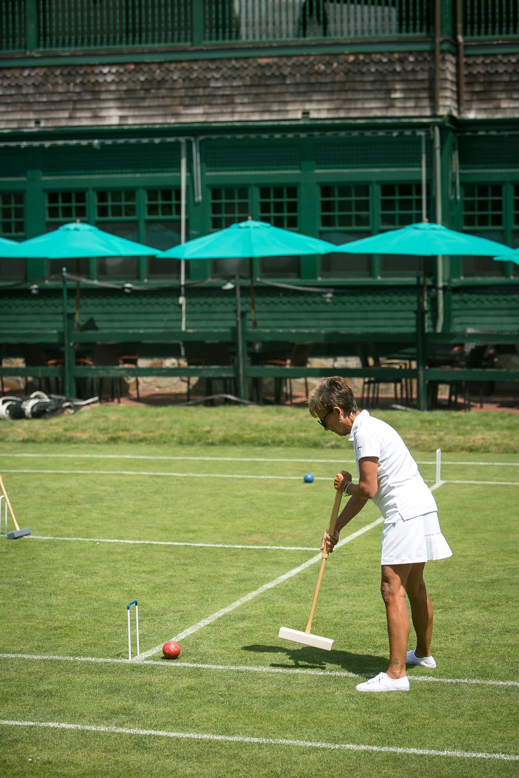 Woman in white playing croquet on a green lawn, green umbrellas and building in the background.