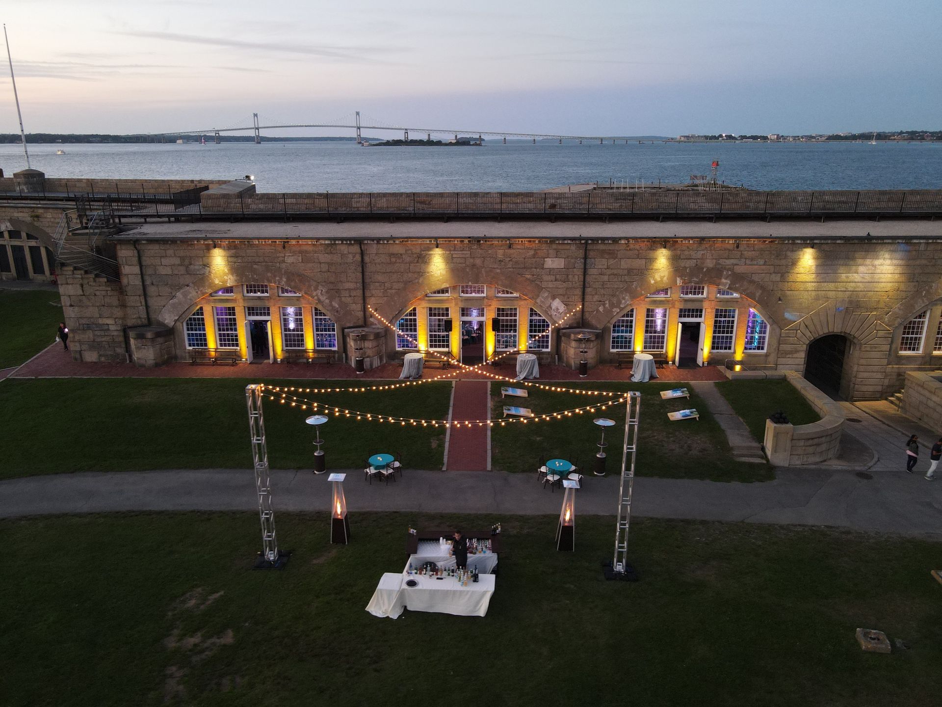 Outdoor event at a stone building near water, with lights strung overhead. A bridge is visible in the background.