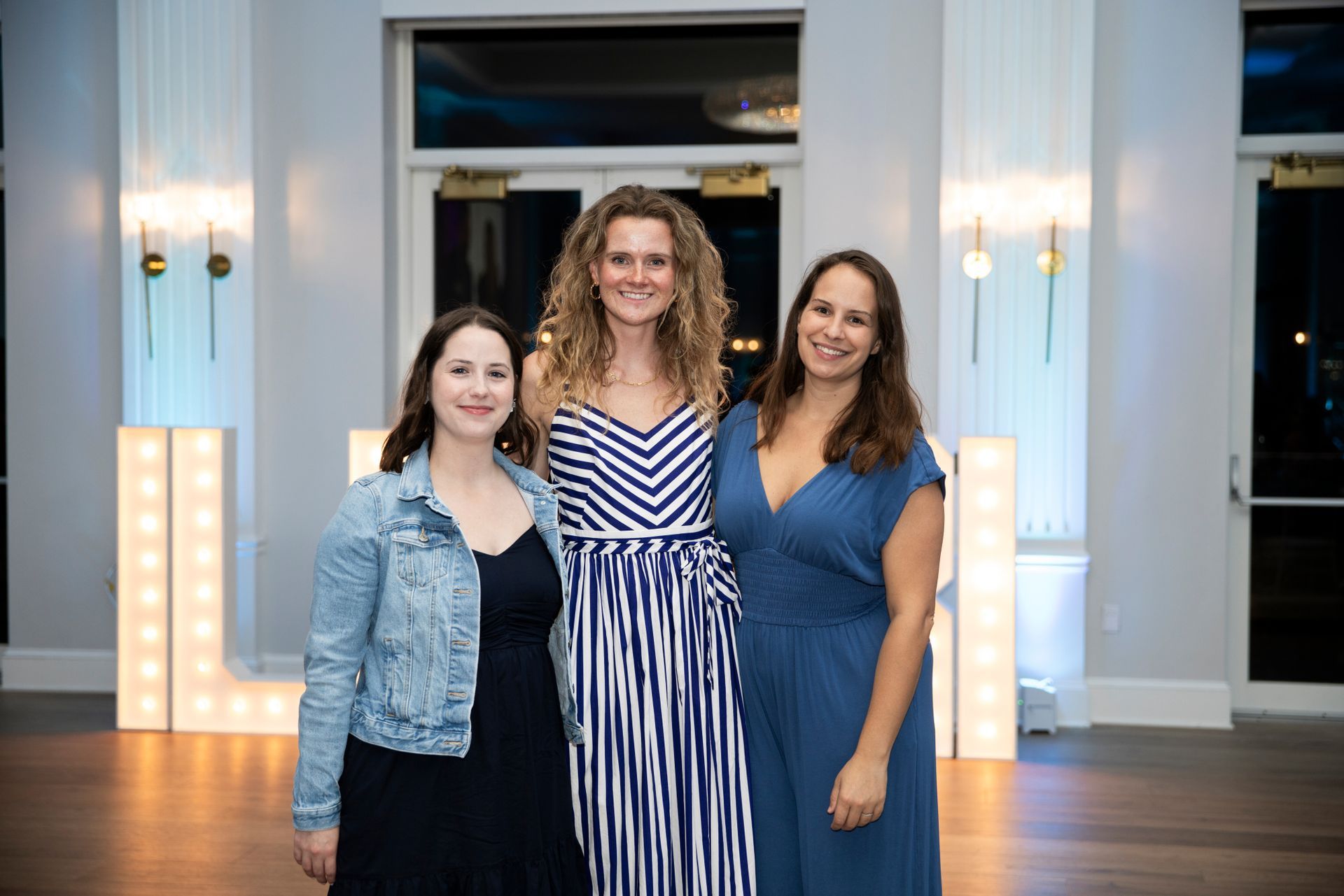 Three women pose in front of illuminated letters. One in a blue dress, one in a striped jumpsuit, and one in a jean jacket.