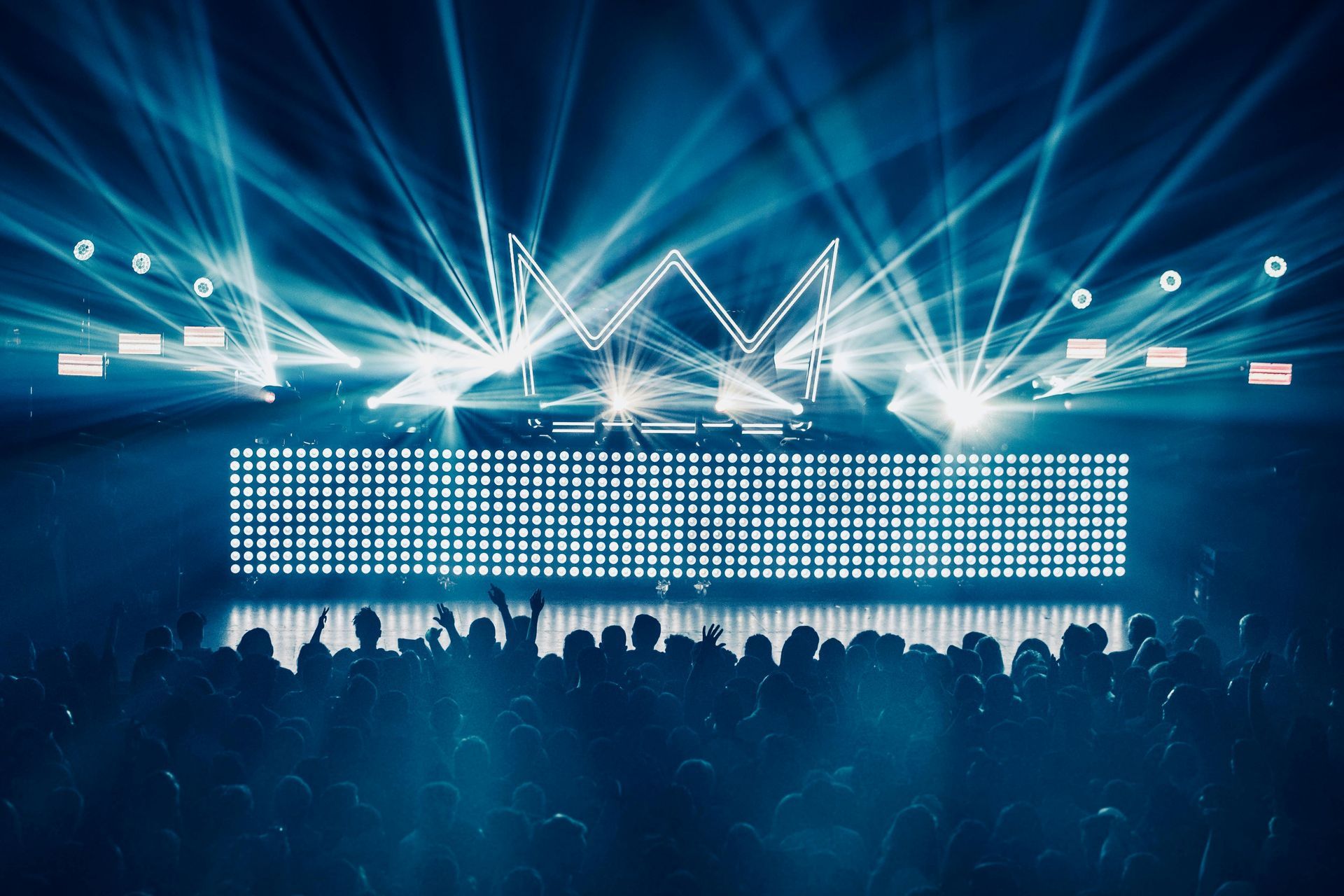 Concert stage with bright blue lights, a crown-shaped structure, and a crowd silhouetted in front.