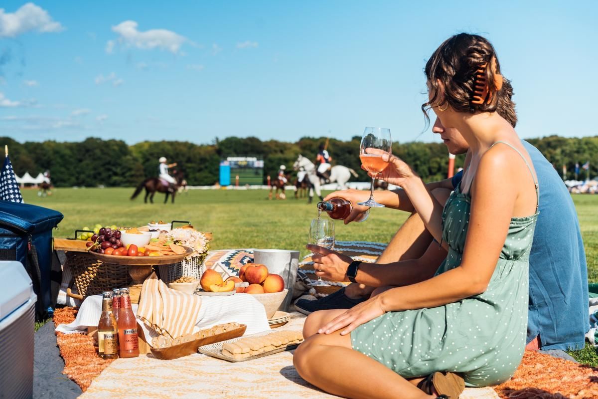 Couple picnicking with food and wine on the grass, watching horses in a competition.