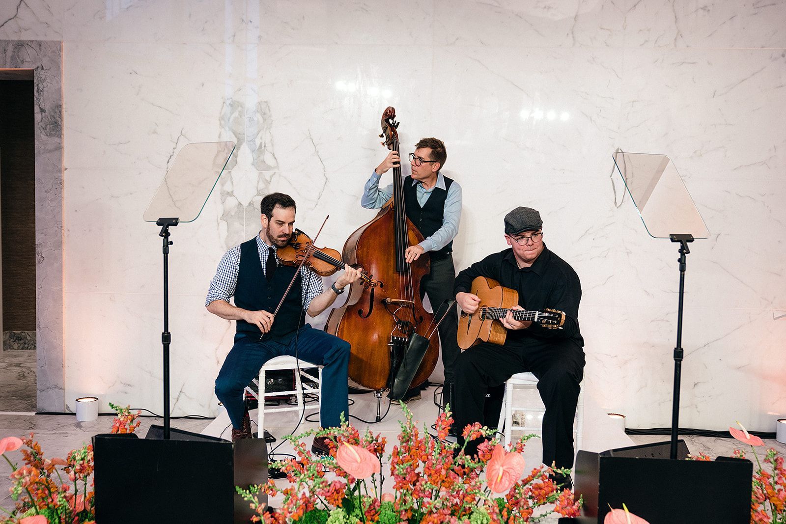 Three musicians play instruments on a stage. One plays violin, one plays bass, and one plays guitar.