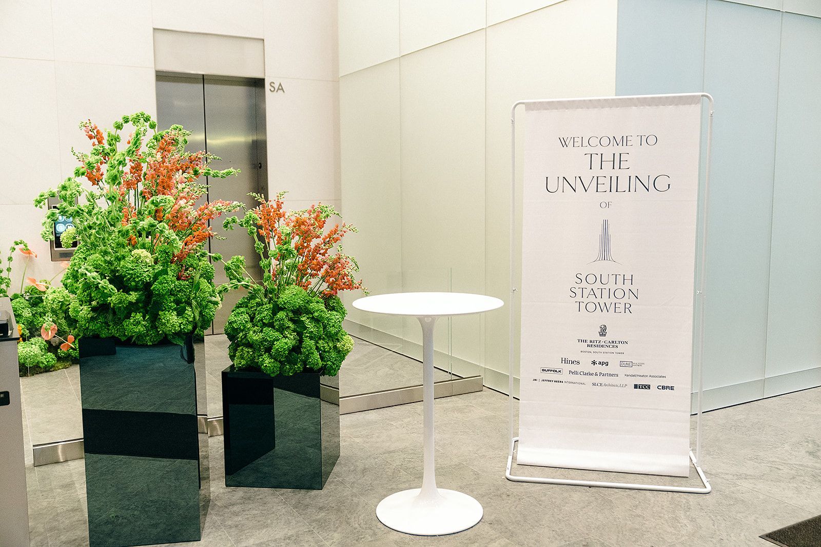 Entrance decorated for an event, featuring a sign, small table, and potted plants next to an elevator.